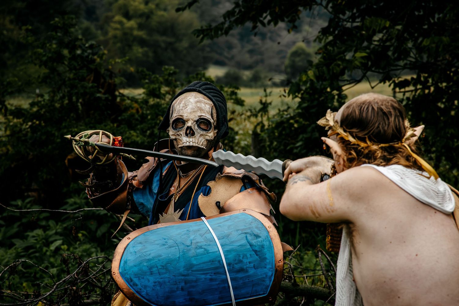 Two costumed performers in a forest, one wearing a skull mask with a blue shield, facing another in a fantasy battle scene.