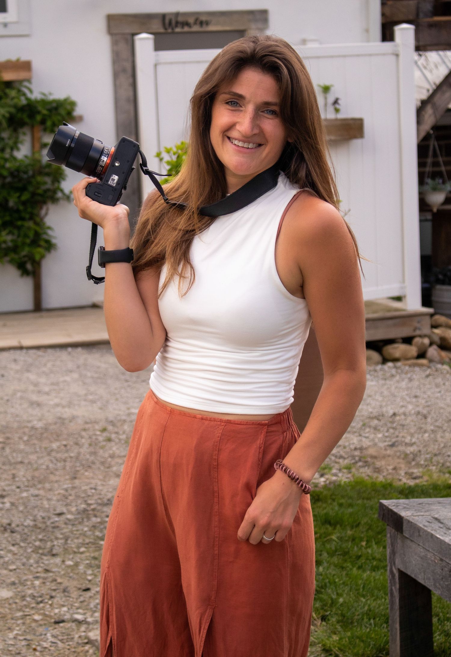 A photographer in a white tank top and rust-colored pants poses with a camera in an outdoor setting.