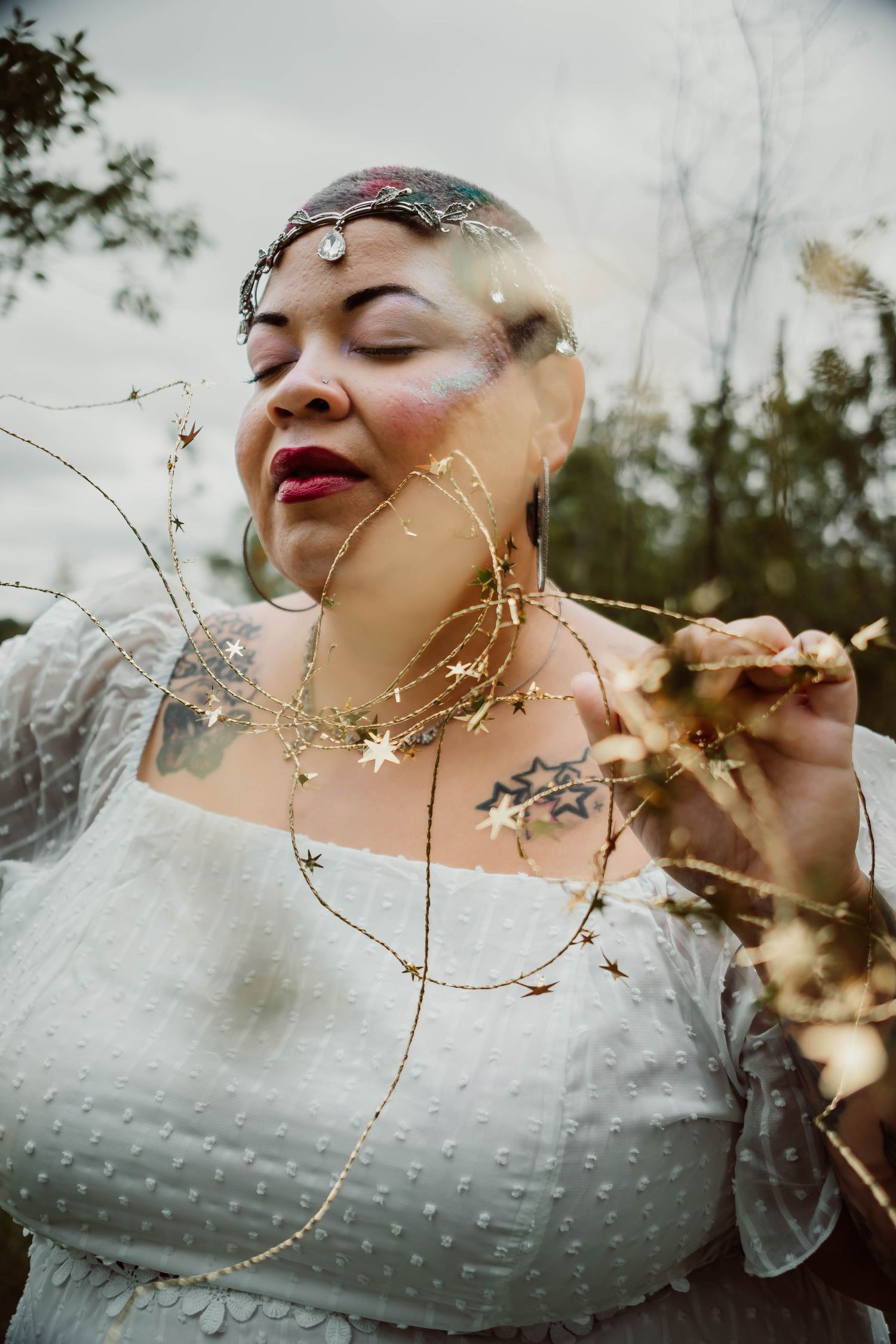 Woman in white dress holding gold star garland, wearing celestial headpiece and star tattoos, outdoors with dreamy double exposure effect.