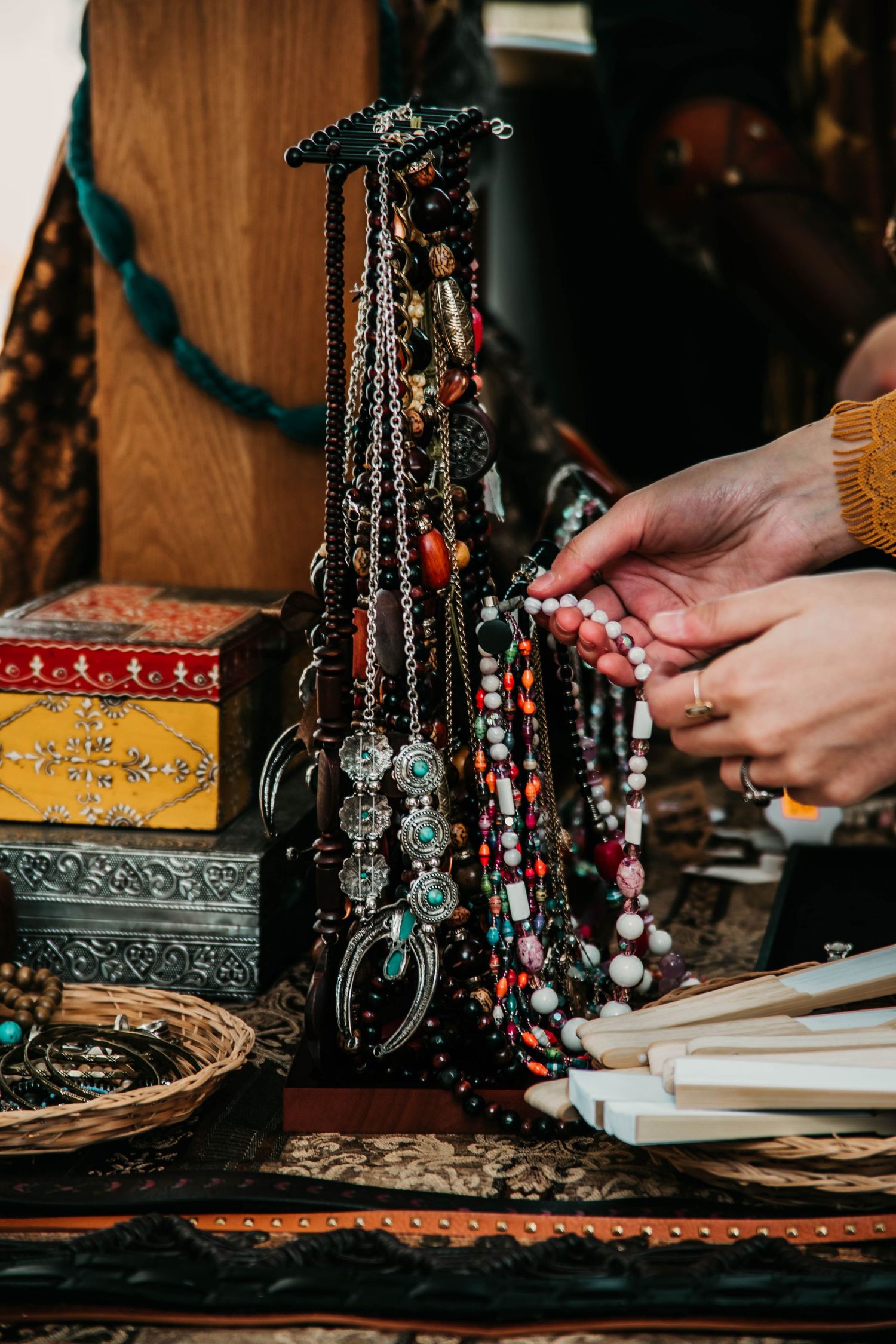 Hands arranging jewelry and beaded necklaces on a rustic wooden surface with ornate boxes in warm lighting.