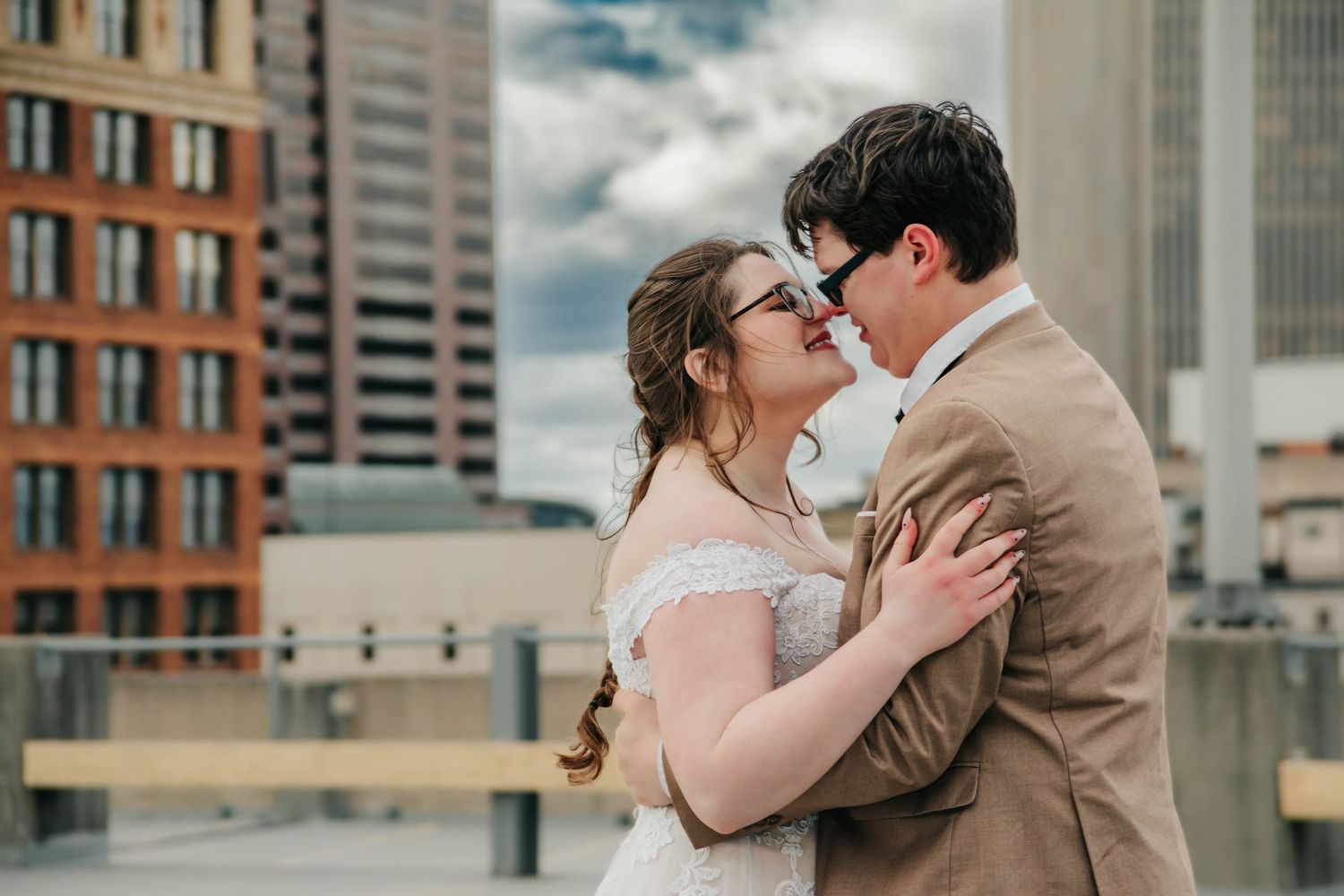 A romantic wedding couple embraces on a rooftop with urban architecture and cloudy skies in the background.