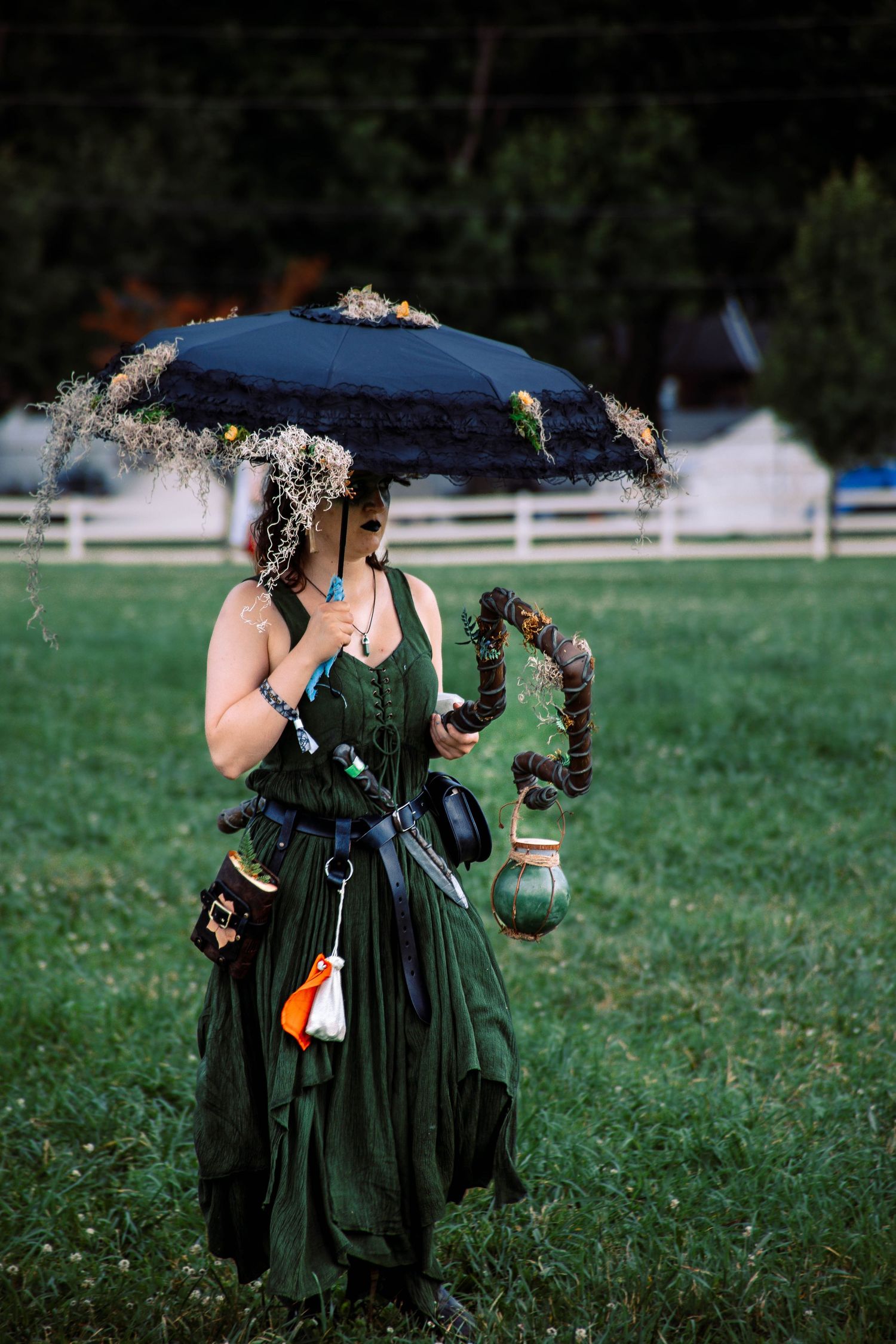 A person in a medieval renaissance faire costume wearing a green dress and holding a parasol stands in a grassy field.