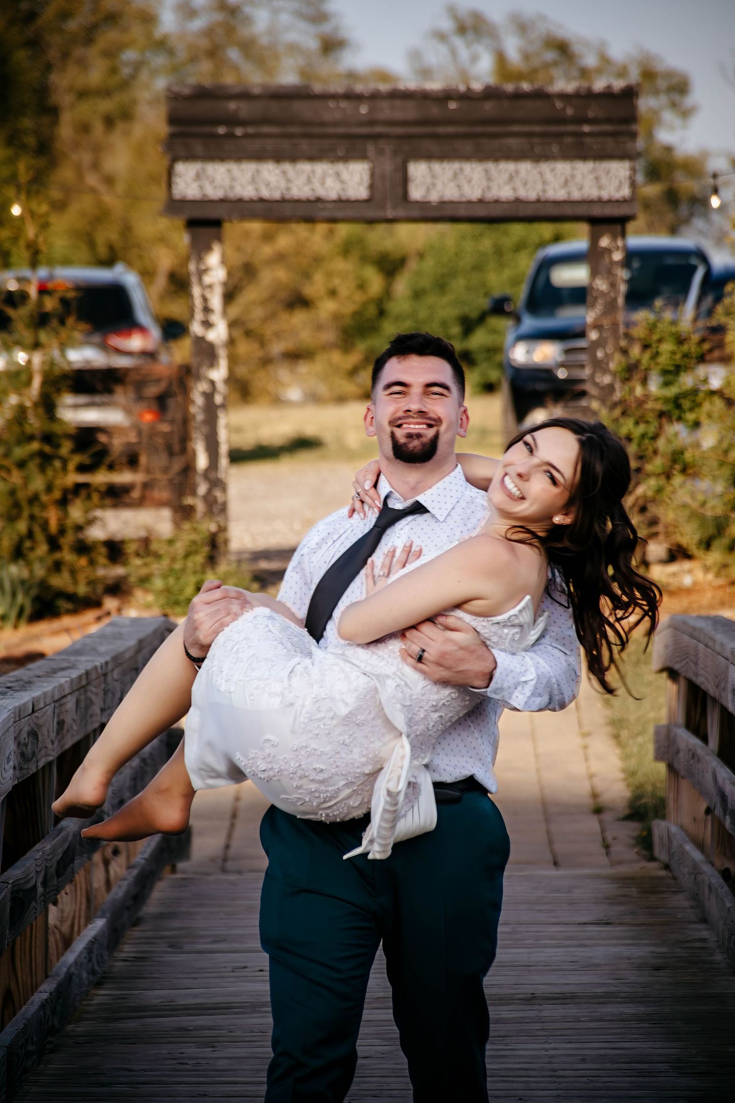 A bride in a flowing white wedding dress twirls playfully while walking with her partner in a wooded outdoor setting.