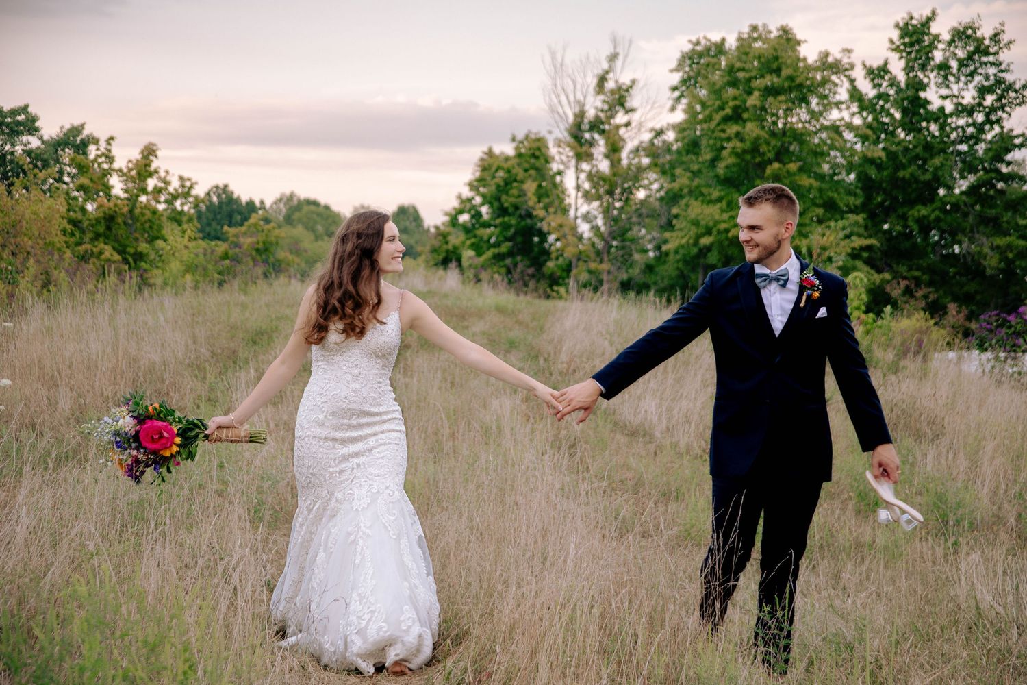 Playful wedding couple running through a meadow at sunset.