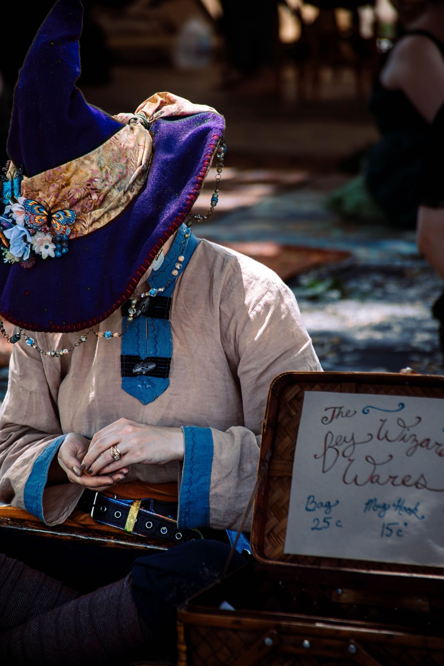 Person wearing a decorative blue hat with crystals and white medieval costume sits at a table with a price sign.