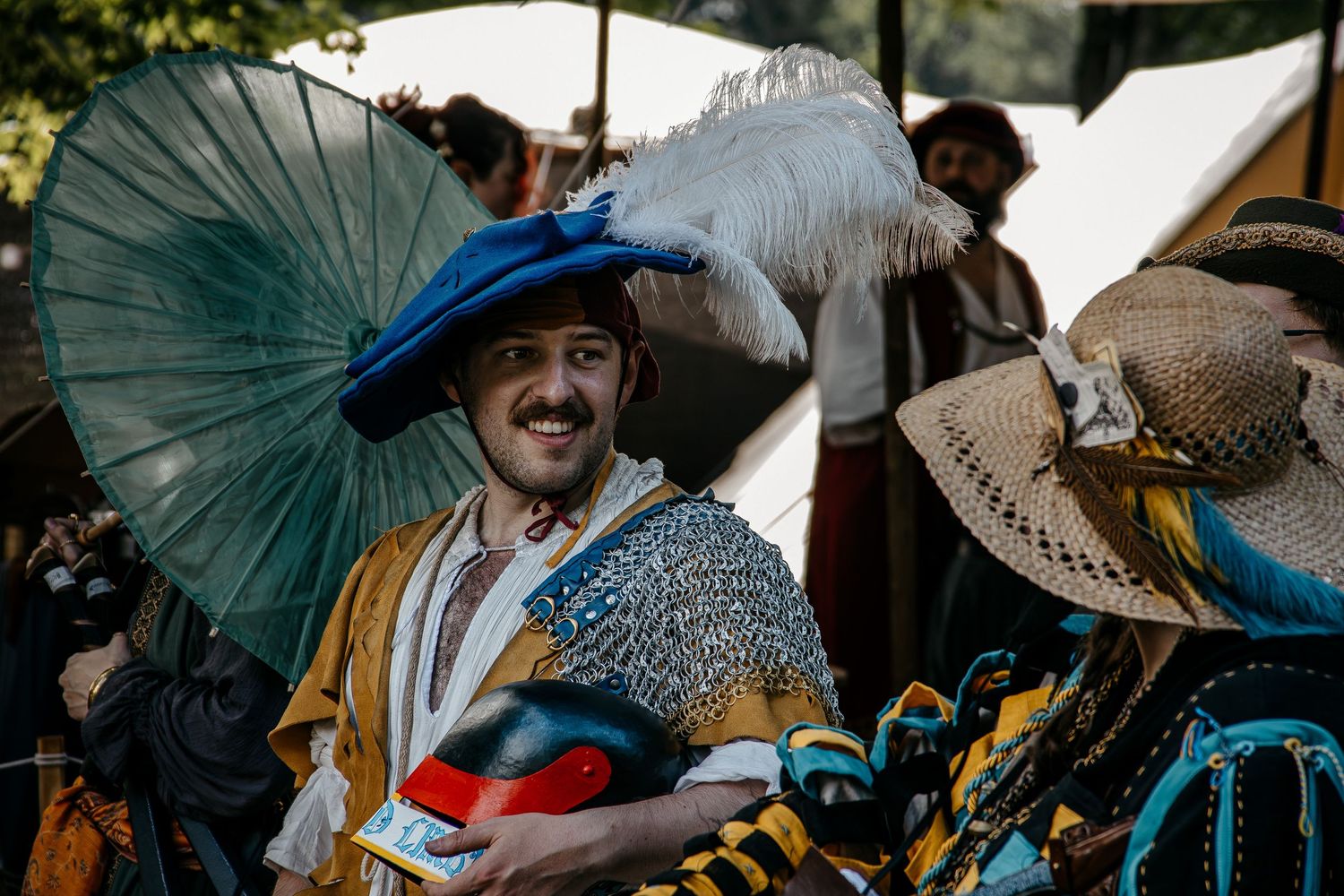 Man in colorful Renaissance festival costume with feathered hat and chainmail smiles at outdoor medieval fair.