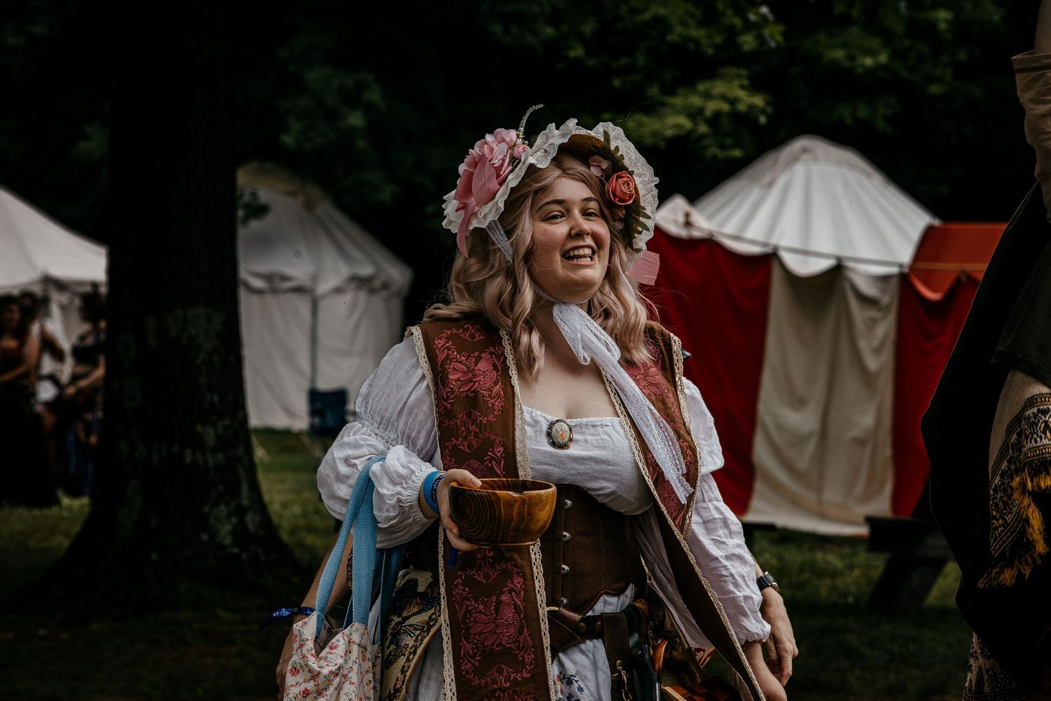 Woman in Renaissance faire costume with floral crown and ornate dress laughs joyfully outdoors near medieval tents.
