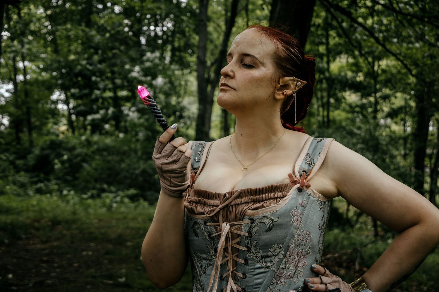 Woman in elf costume with pointed ears holds a pink flower in a forest setting.