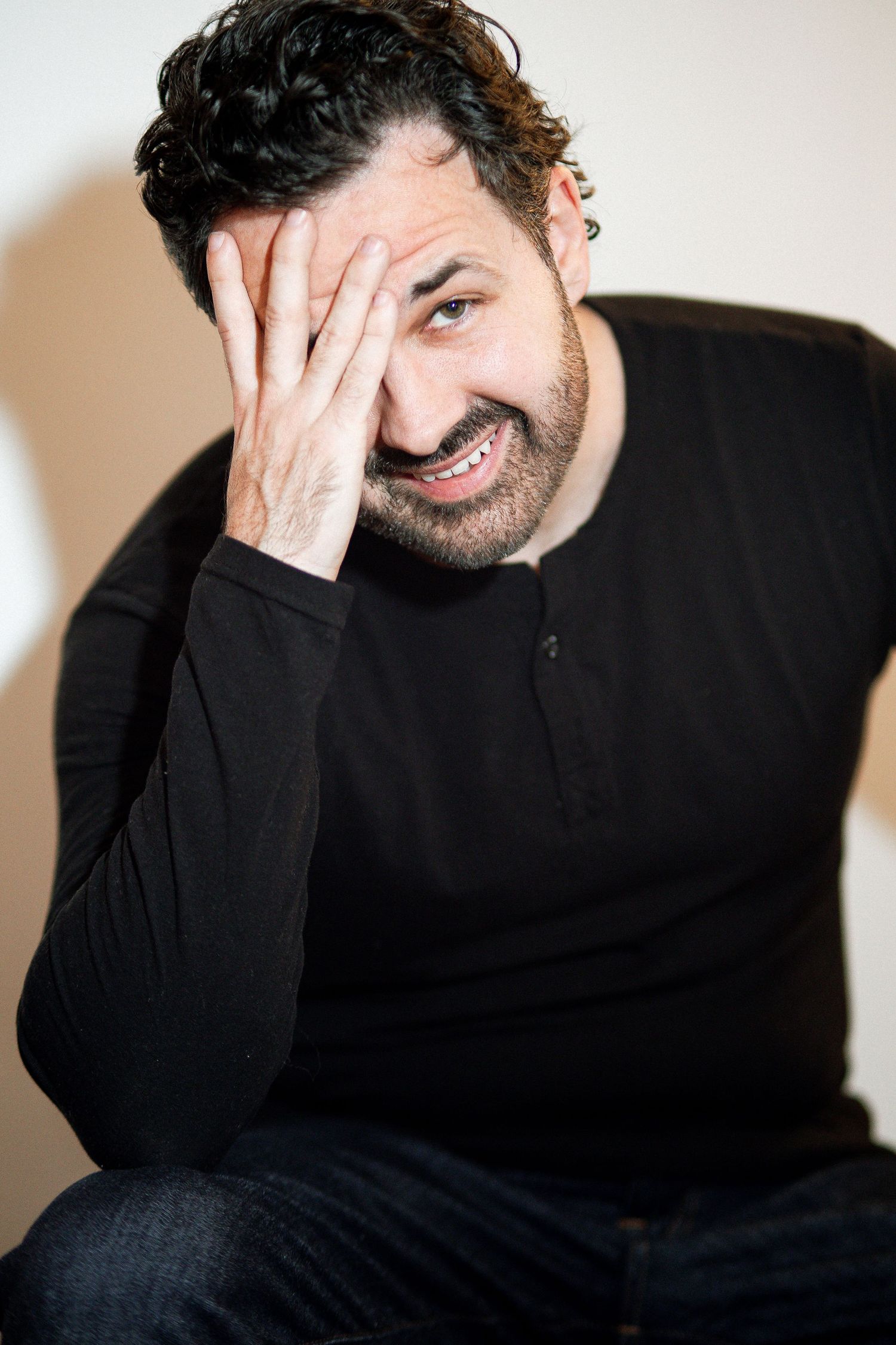 A bearded man in a black long-sleeve shirt sits smiling with his hand on his face in a casual portrait.