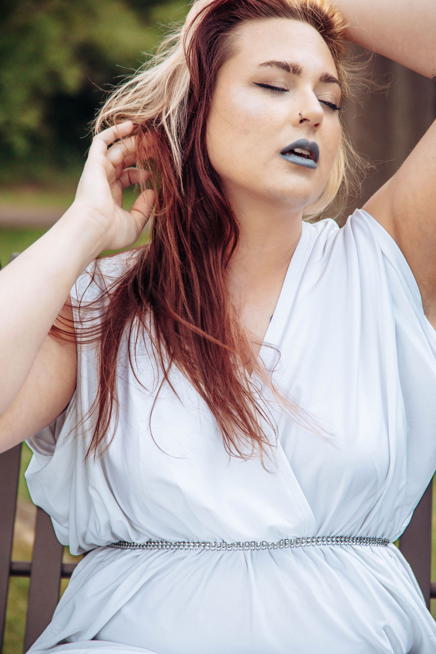 A plus-size woman with red hair in a white dress and dark lipstick poses outdoors with eyes closed.