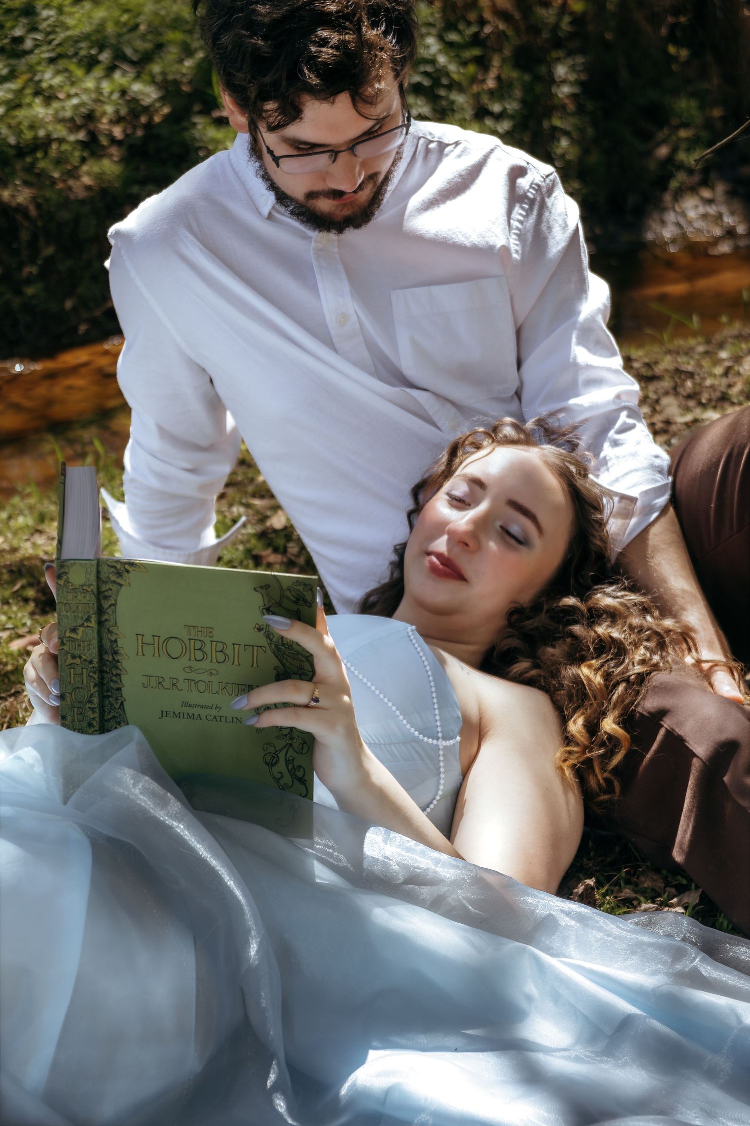 Bride in white gown reads The Hobbit outdoors while groom in white shirt looks over her shoulder.