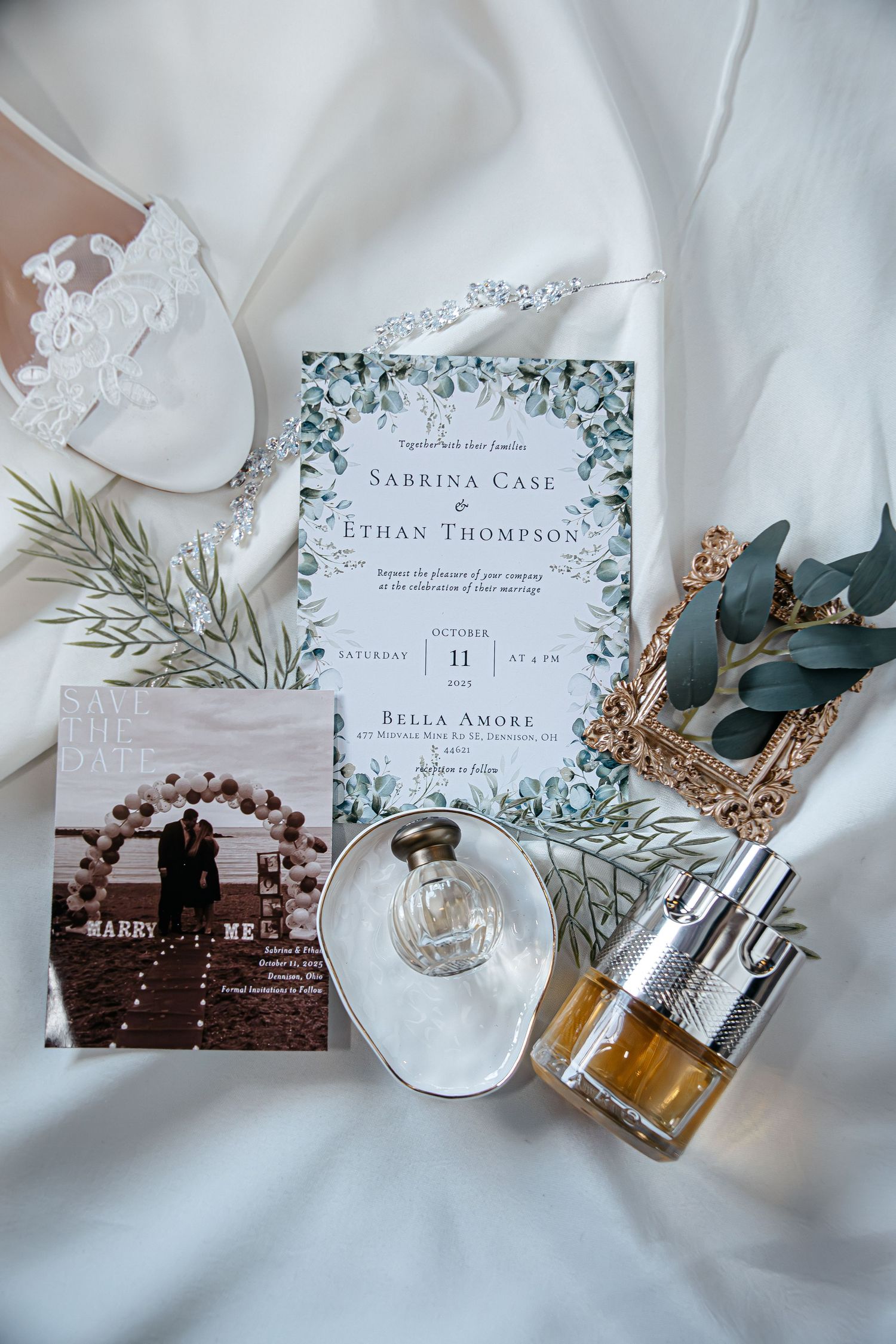 Wedding invitations with floral designs displayed alongside perfume bottles and jewelry on white fabric.