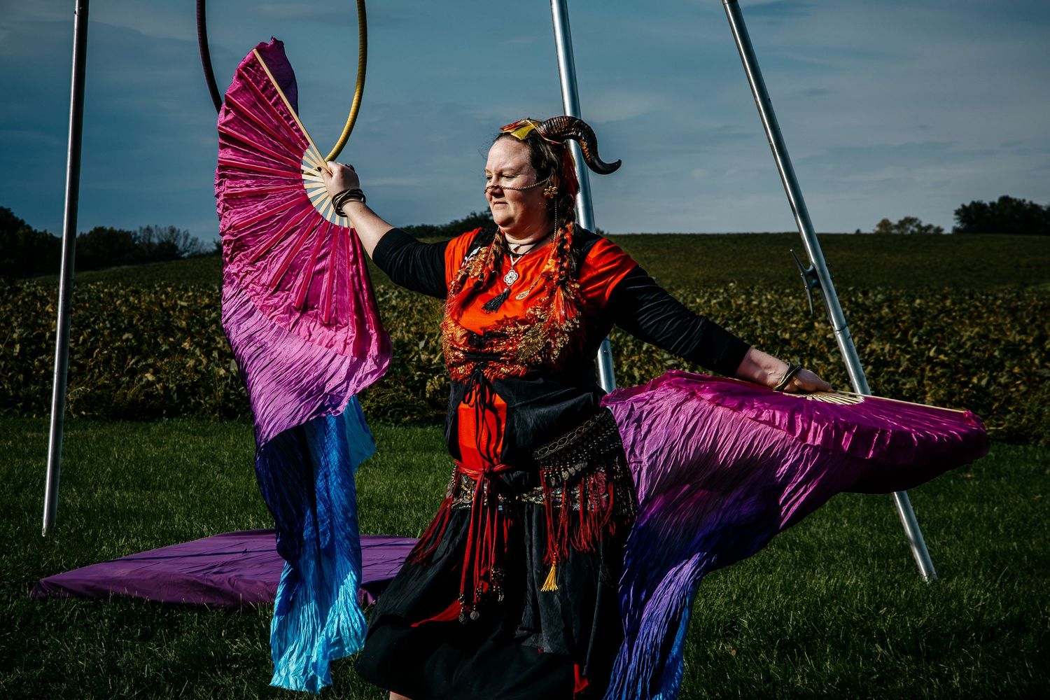 Woman in colorful costume holds vibrant pink and blue silk fans outdoors near aerial hoop equipment.