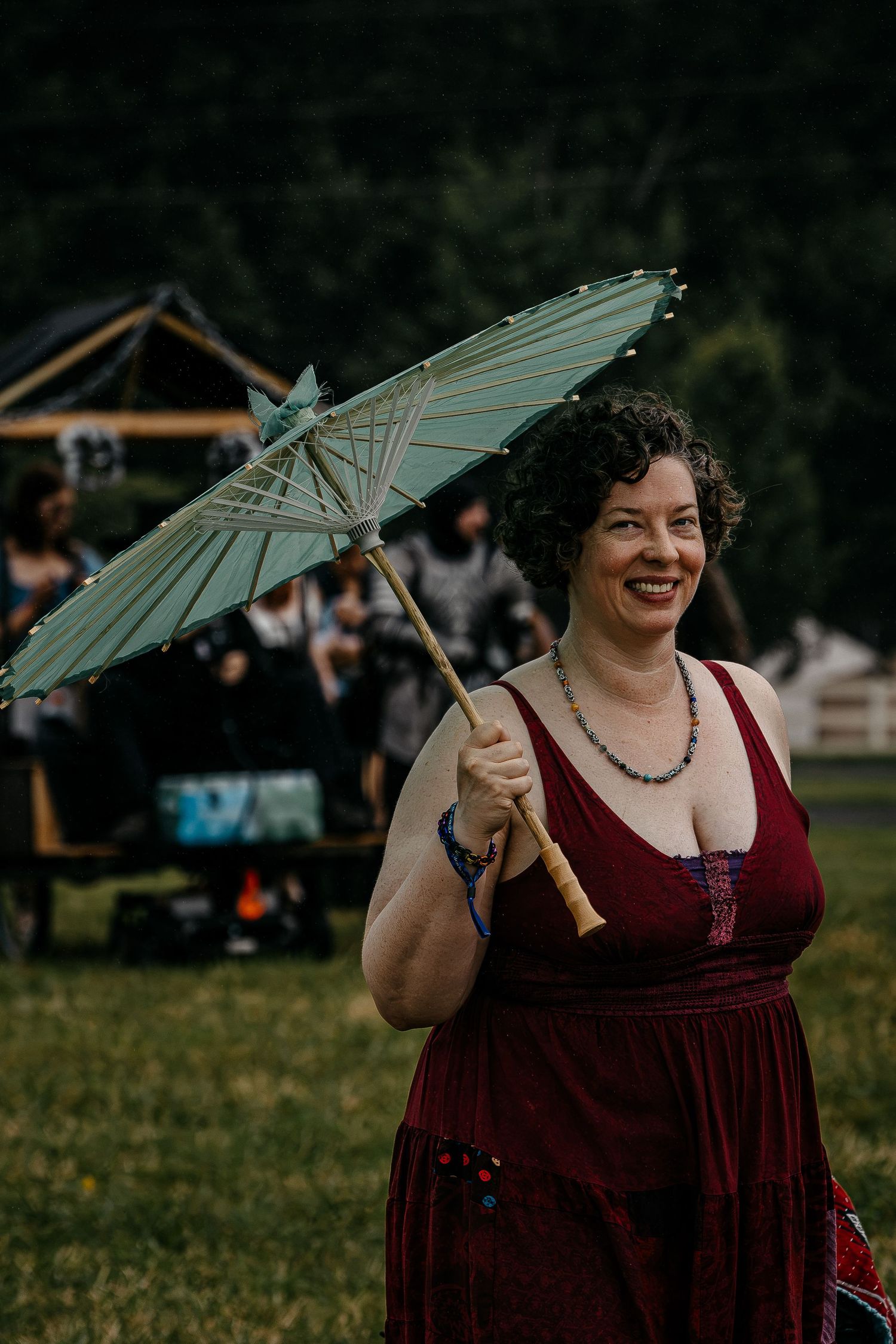 Smiling woman in burgundy dress holding a teal parasol umbrella outdoors at a festive event.