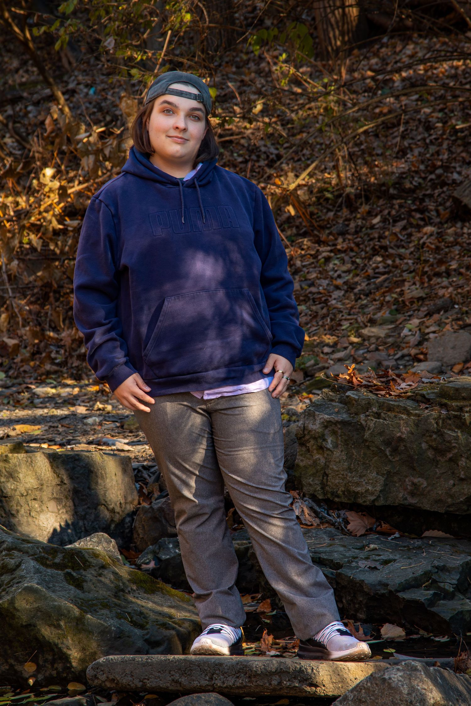 Person in navy hoodie and gray pants sits casually on rocky outcrop in autumn forest setting.
