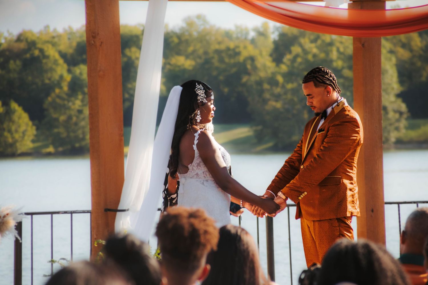 A couple enjoys a romantic moment on a covered deck overlooking a serene lake at sunset.