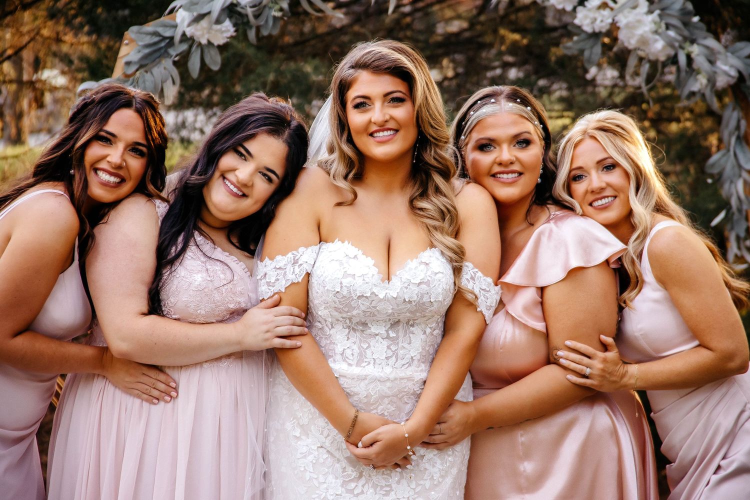 Bridesmaids in pink and blush dresses smile together in a candid outdoor photo shoot.