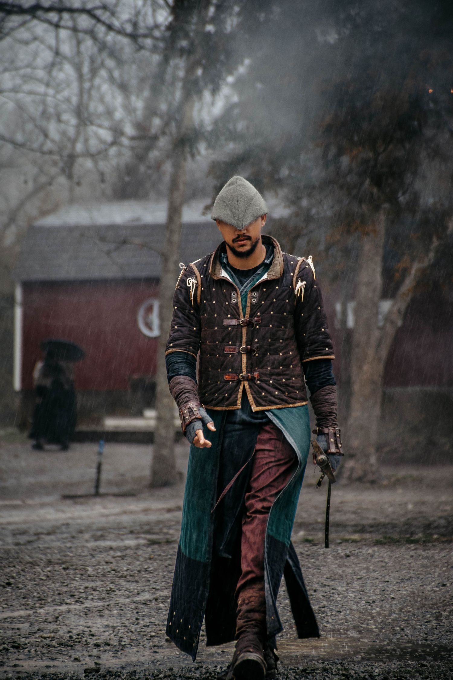 Medieval fantasy cosplay figure in dark armor and cloak stands in misty forest setting with red barn in background.