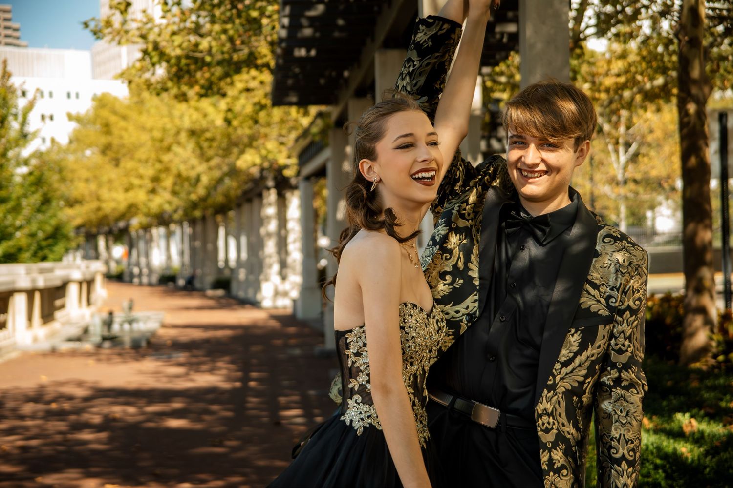Two people in elegant black and gold formal attire laugh together on a tree-lined autumn street.