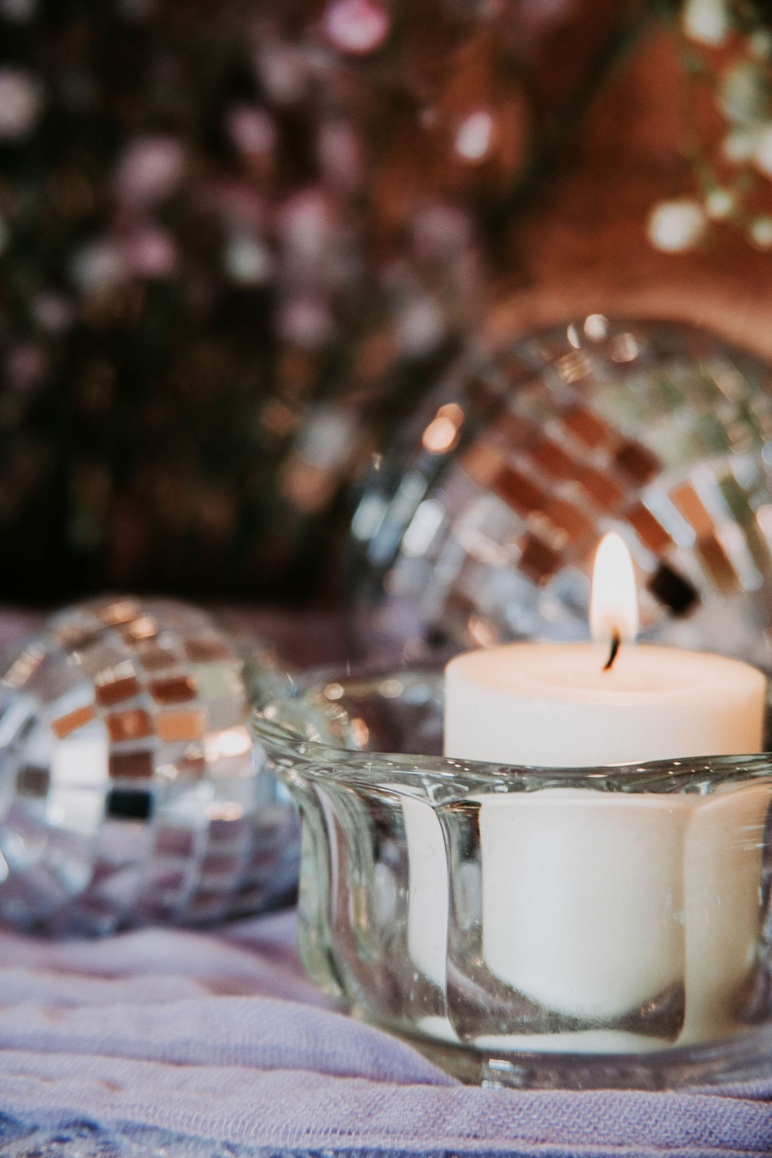 A row of white tea light candles in glass holders on a light fabric surface with bokeh lights in the background.