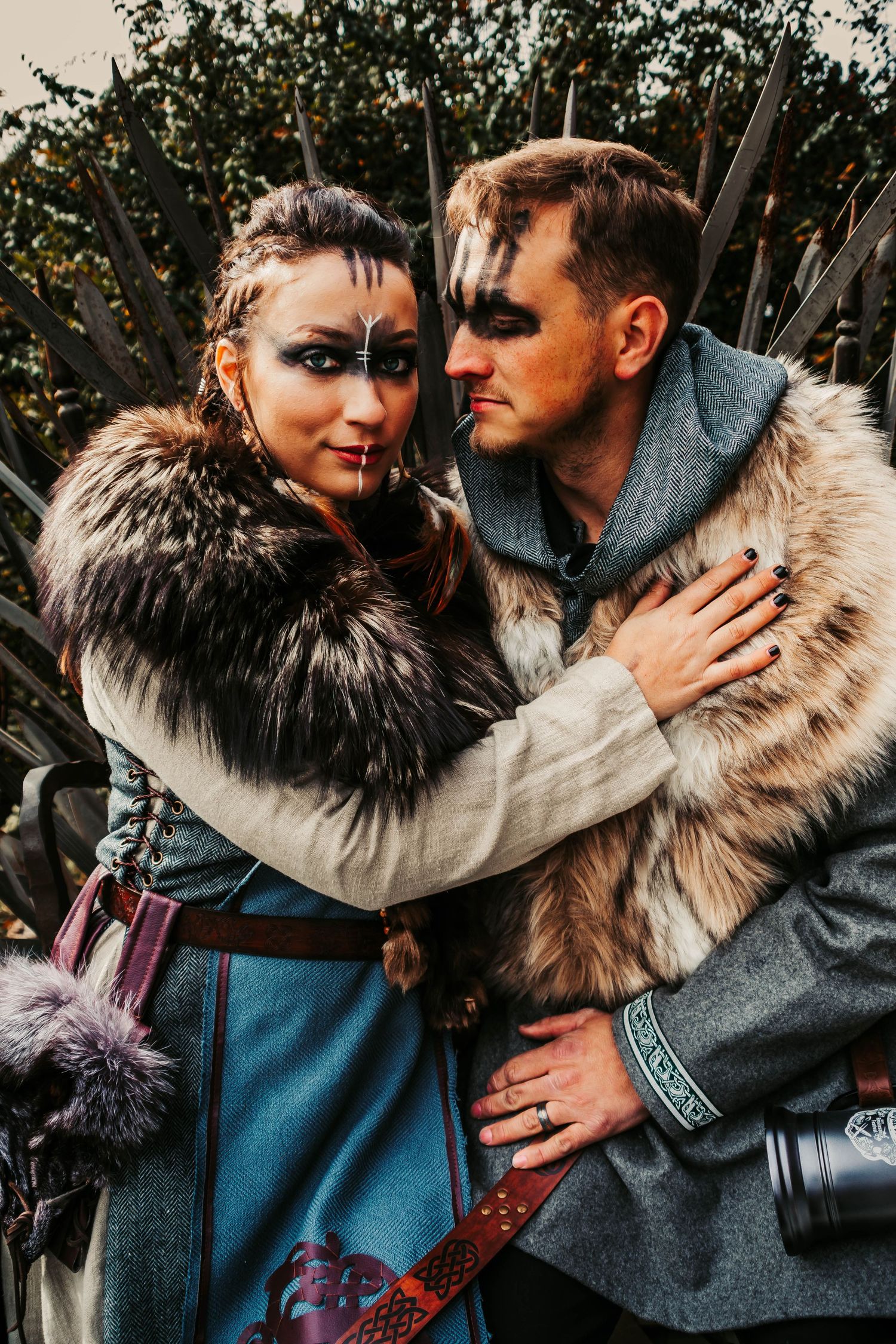 Two people in Viking-inspired costumes with fur collars and dramatic makeup embrace in a moody outdoor setting.