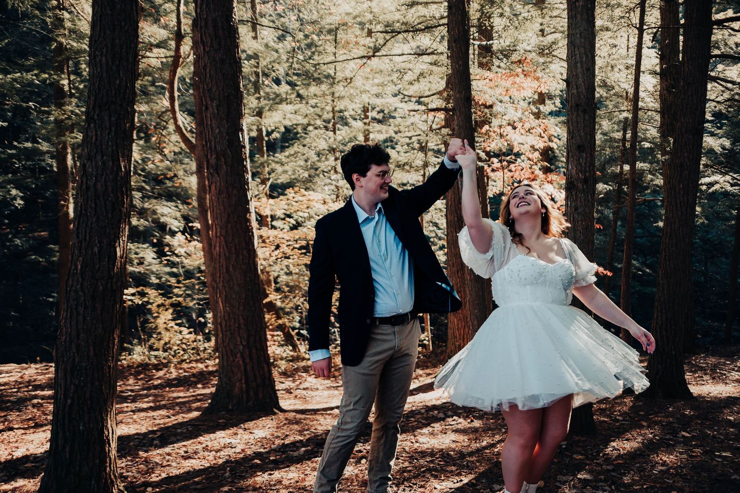 A couple dances joyfully among tall trees in a sunlit forest, the bride in a white dress and groom in a dark blazer.