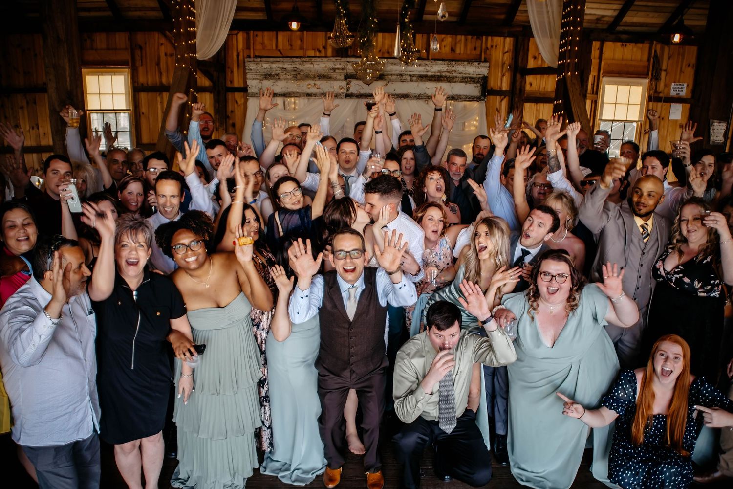 Joyful wedding guests celebrating together in a rustic barn venue, arms raised in excitement during group photo.