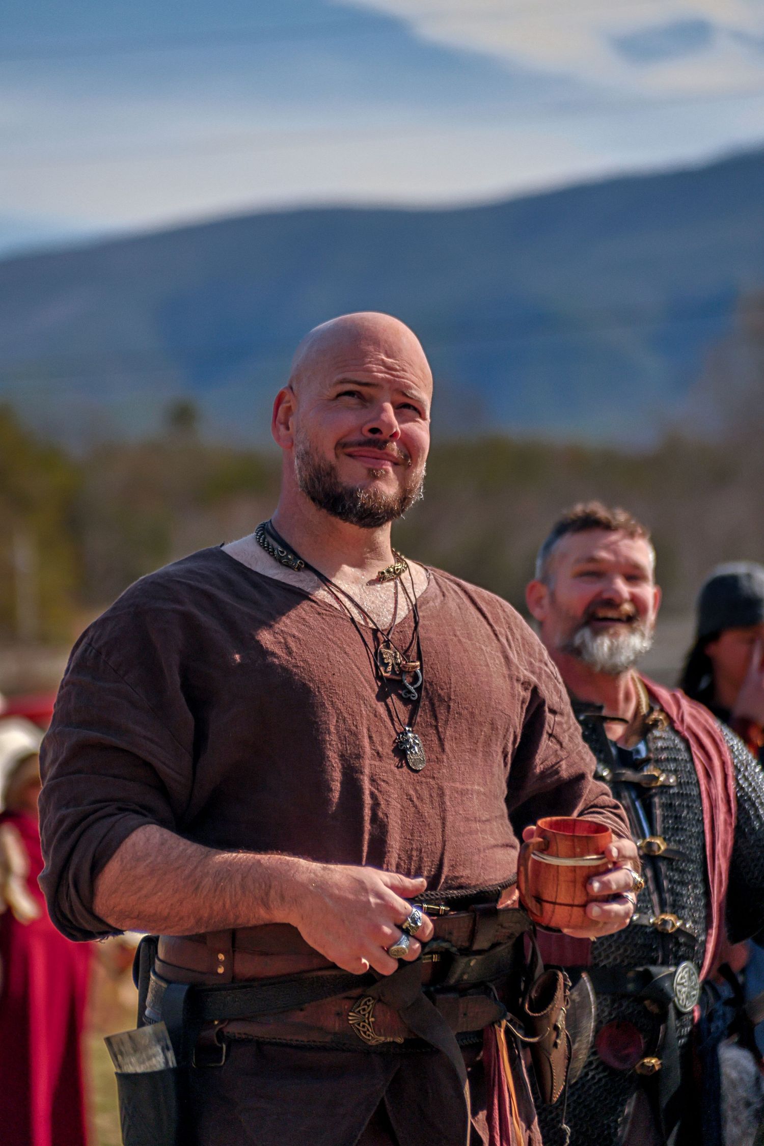Men in medieval costume drink from wooden tankards at a historical reenactment festival with mountains in background.
