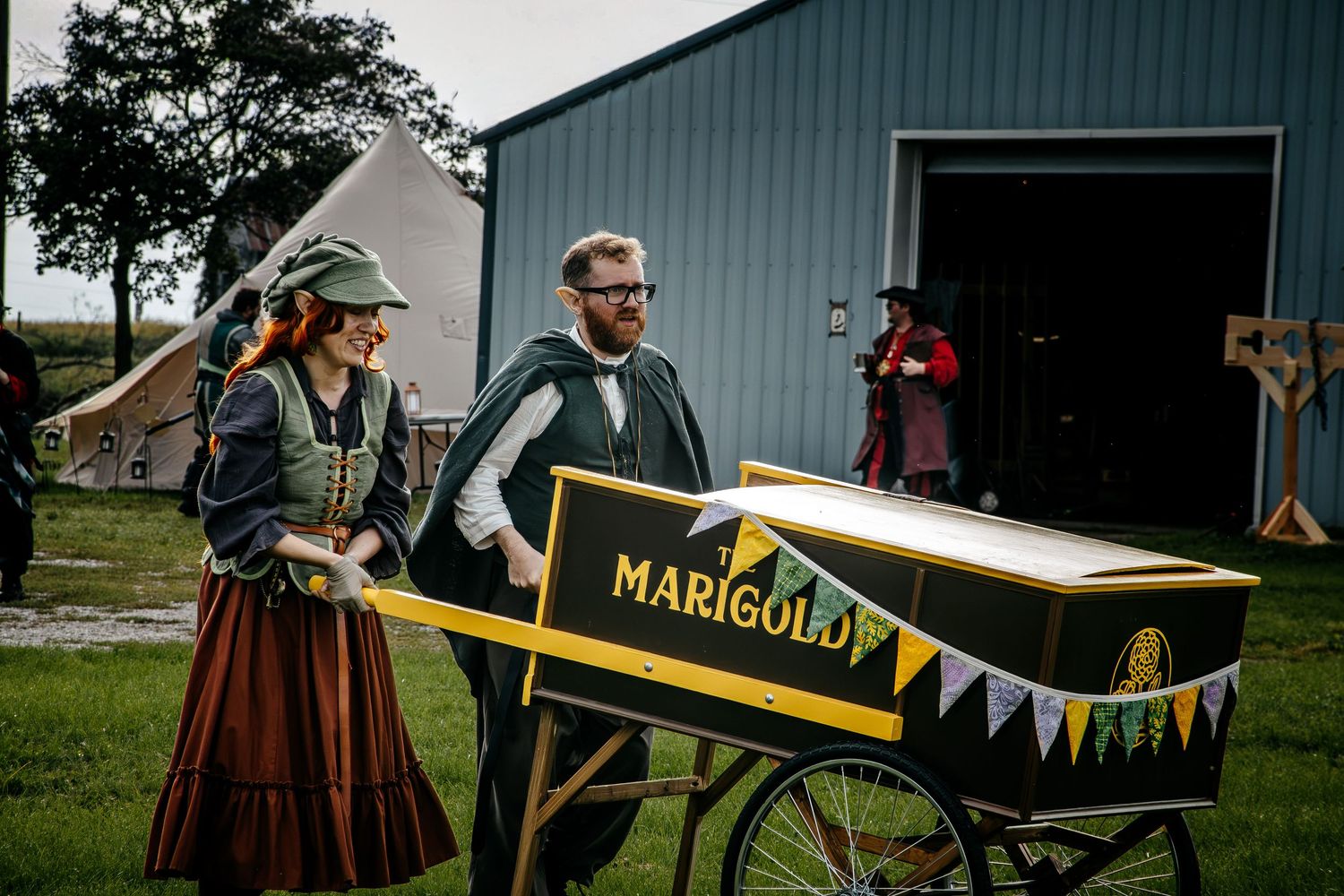 Man and woman in period costumes pushing a vintage Marigold branded cart at an outdoor historical reenactment event.