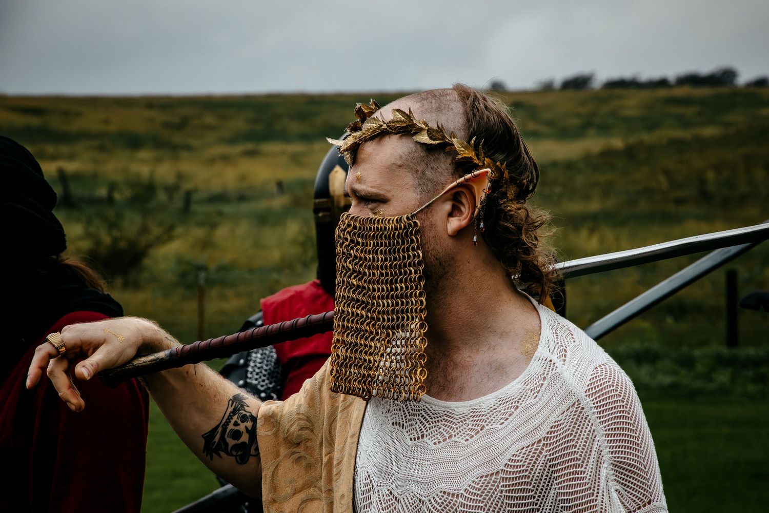 Person in medieval costume with chainmail face guard, pointed elf ears, and laurel crown at outdoor LARP event.