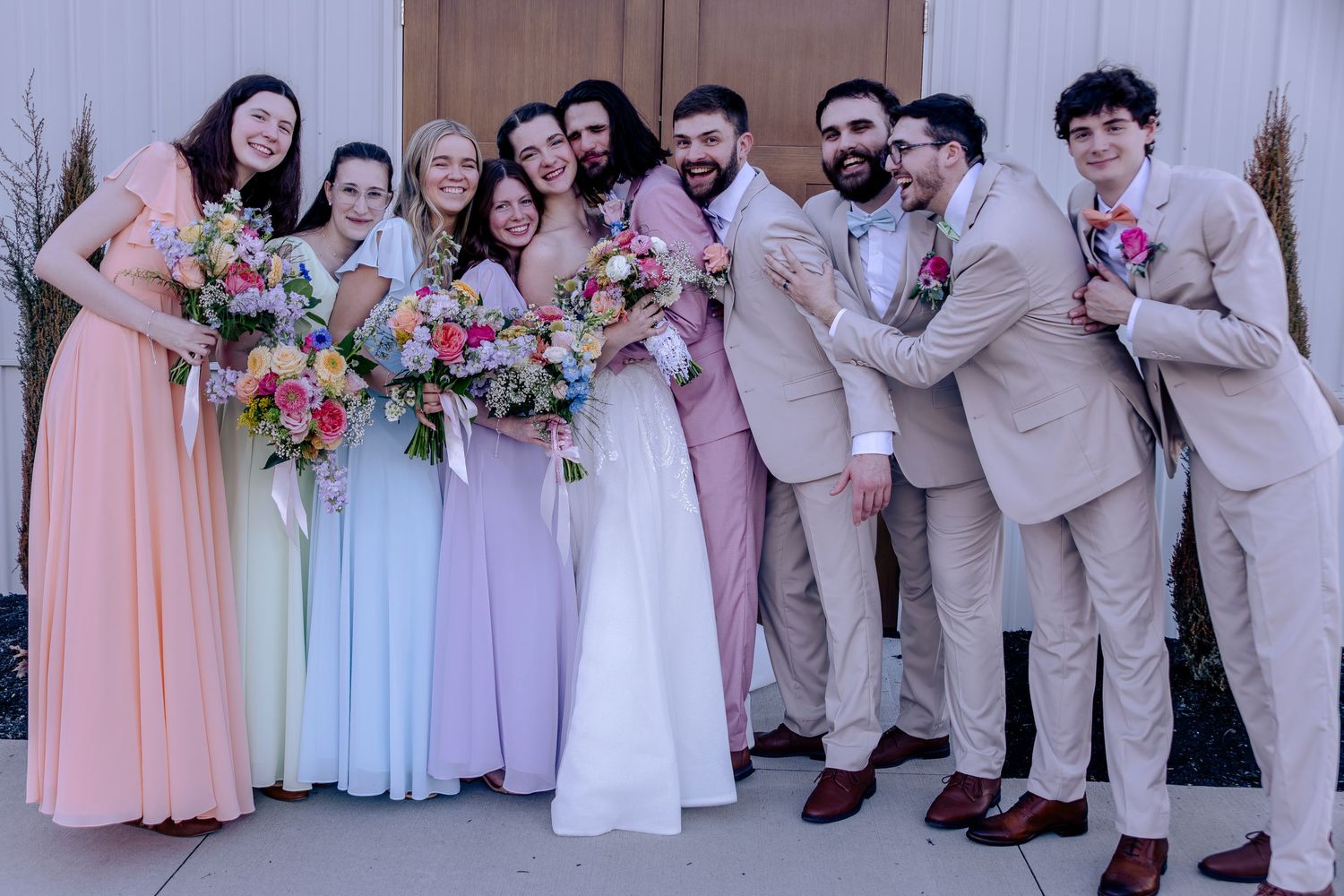 Wedding party poses together, bridesmaids in pastel gowns holding colorful bouquets, groomsmen in tan suits, bride and groom centered.