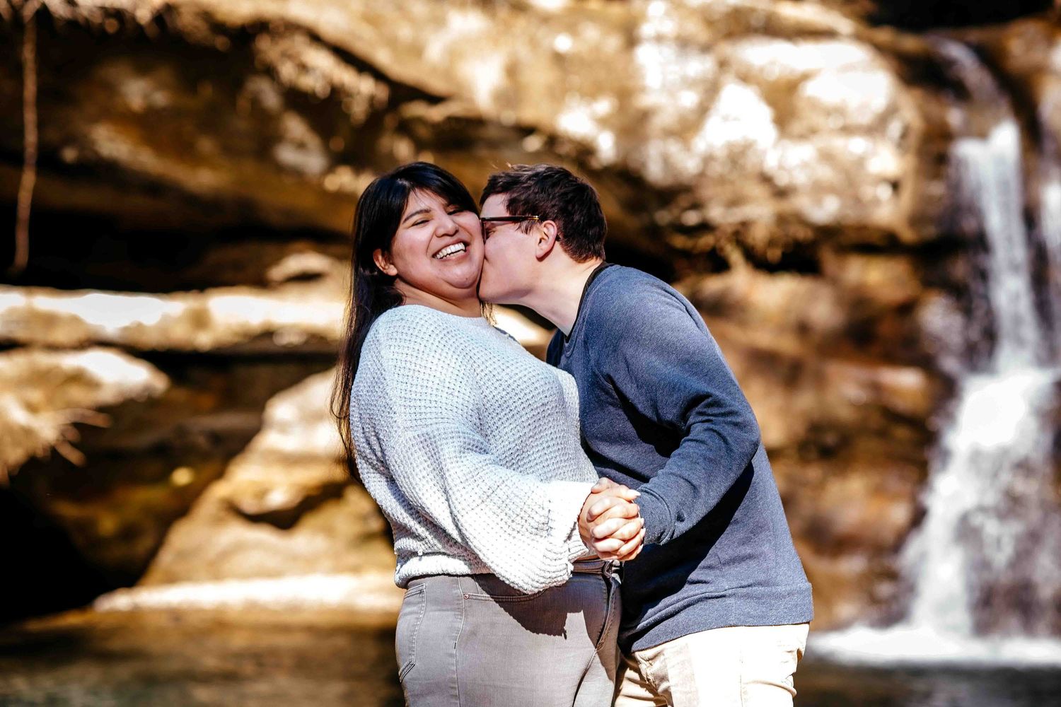 Couple embracing and laughing in front of a waterfall, man kissing woman on the cheek outdoors.