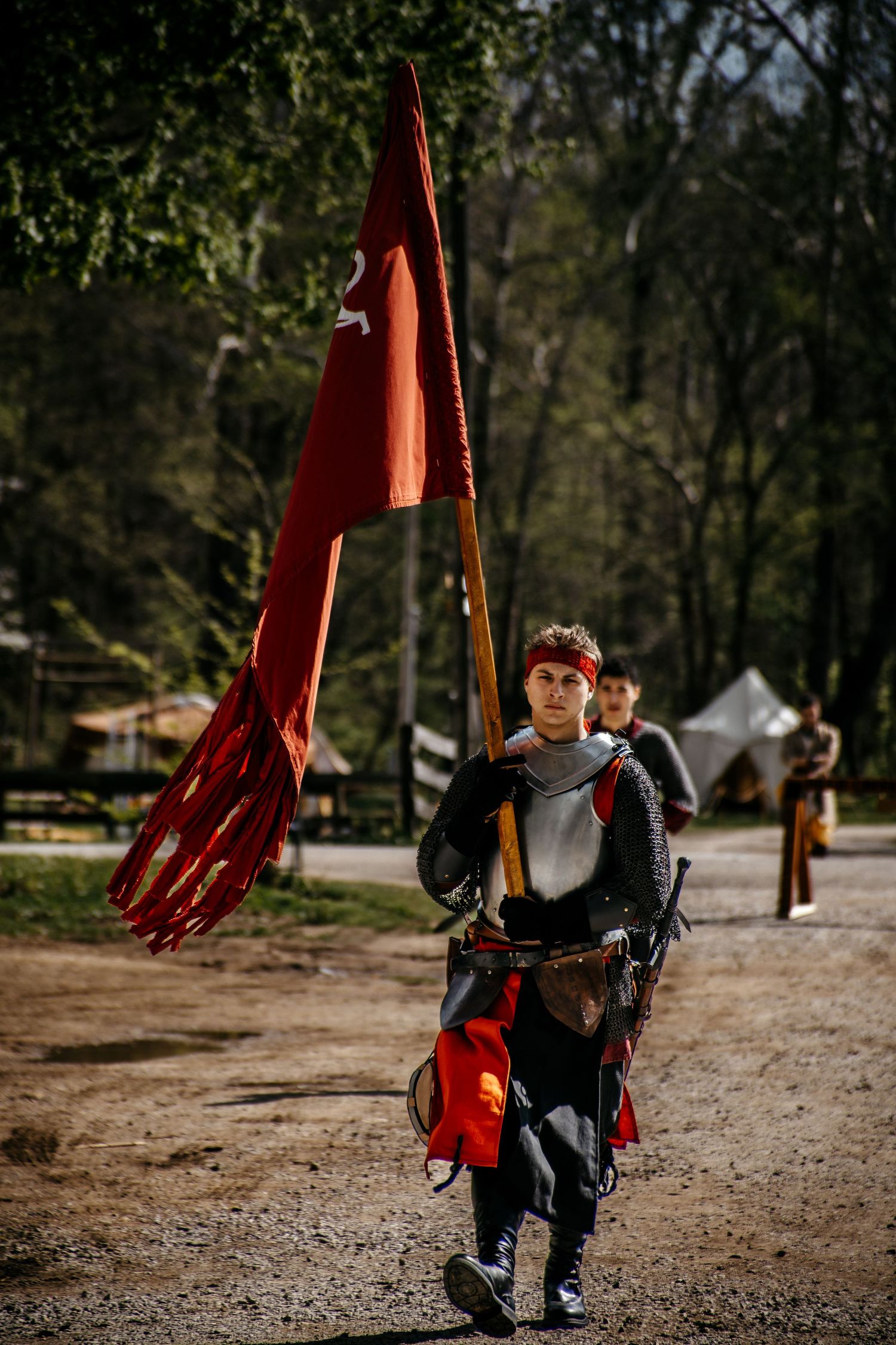 Medieval knight in armor carrying a red flag walks along a forest path during a historical reenactment event.