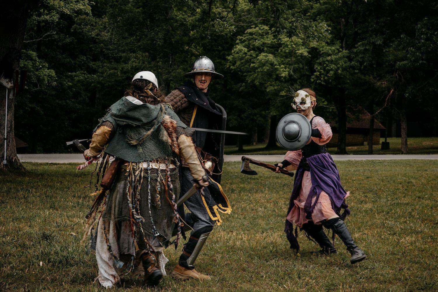 Three people in medieval armor reenact a battle scene outdoors, wielding swords and shields on a grassy field.