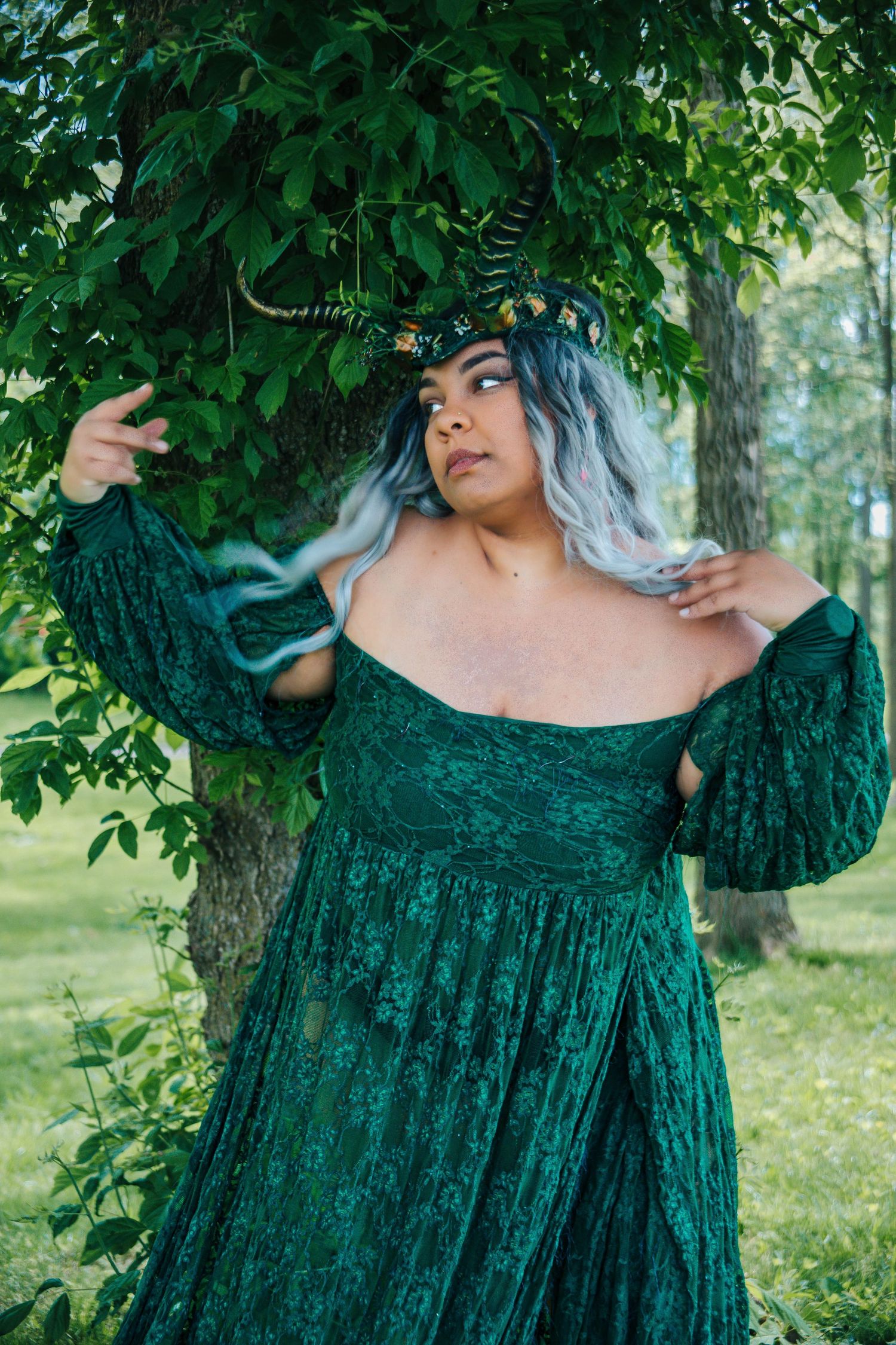 Woman in green lace off-shoulder gown and horned crown poses dramatically by a tree in an outdoor fantasy photoshoot.