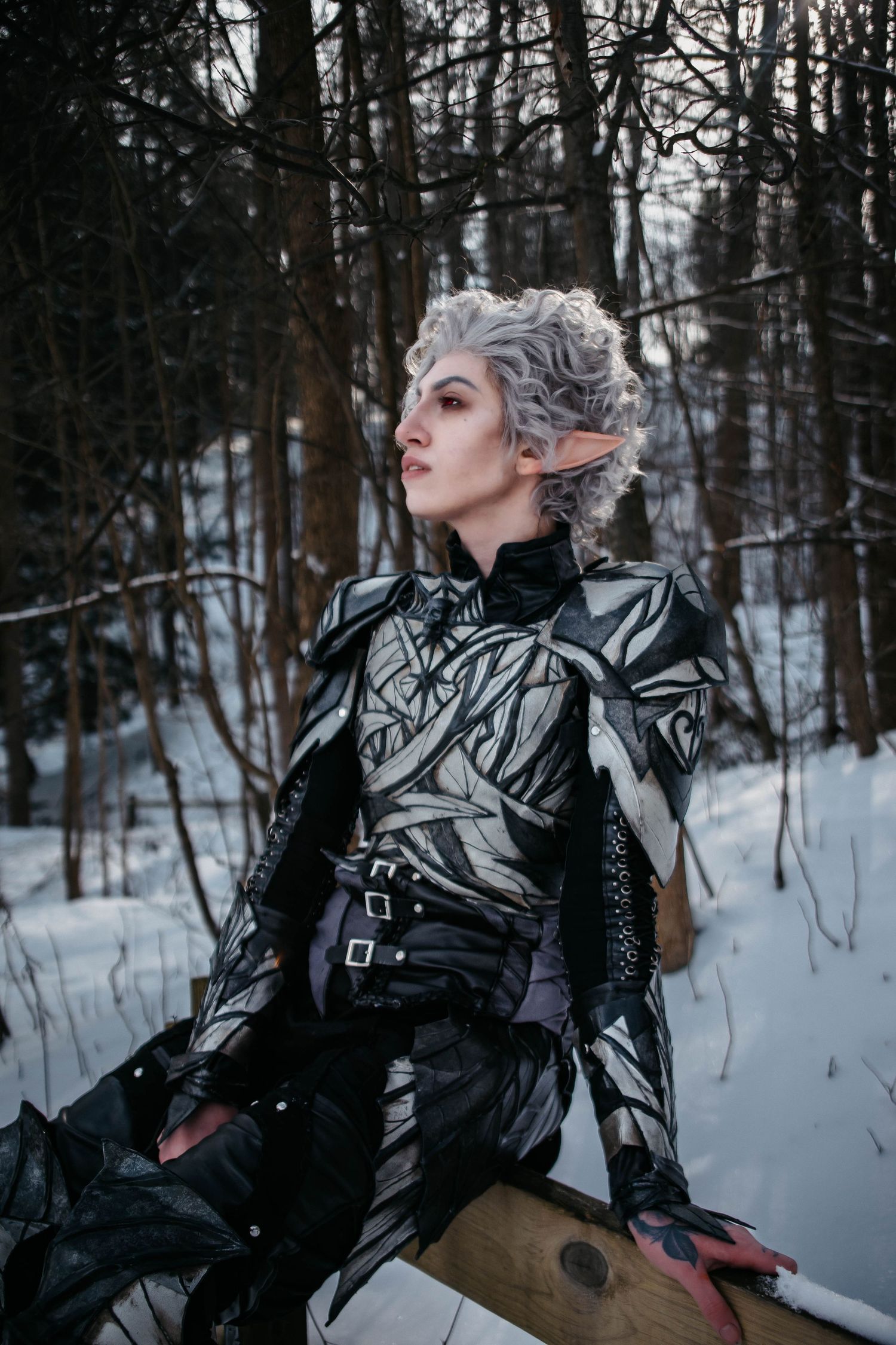 Fantasy cosplay character in silver armor and white hair poses dramatically in a snowy winter forest setting.