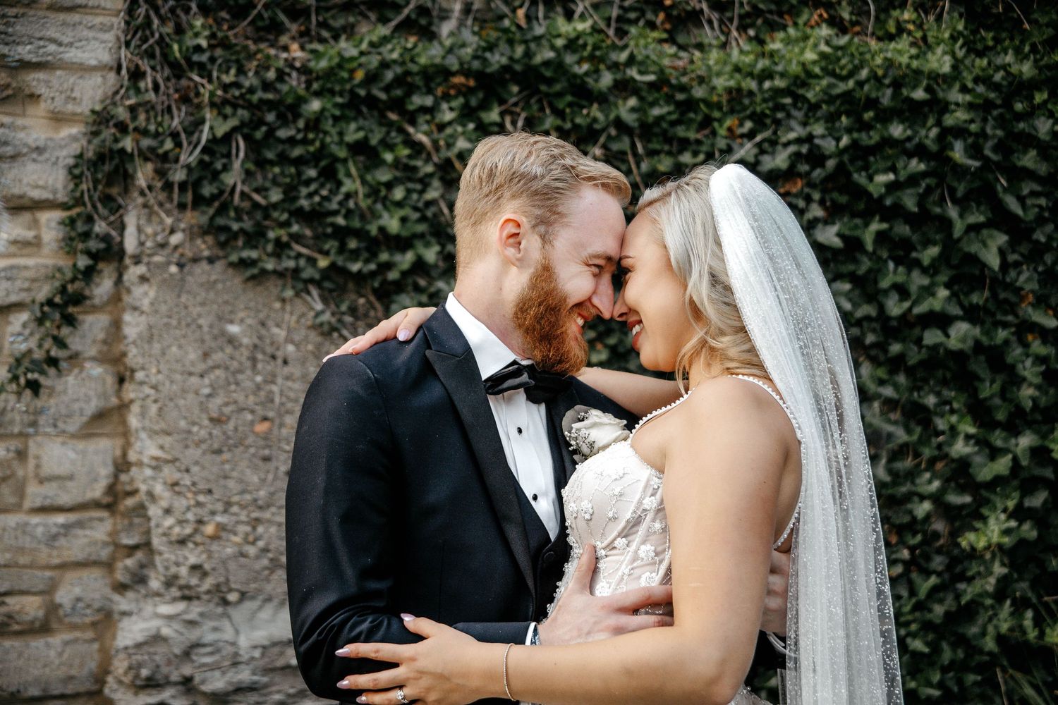 Romantic wedding moment captured against ivy-covered stone wall.