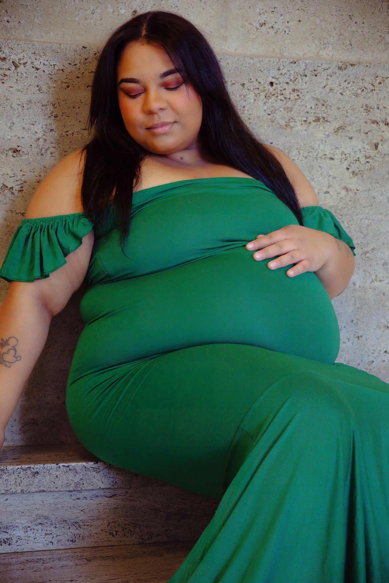 A maternity portrait featuring an elegant green off-shoulder dress against a textured wall.
