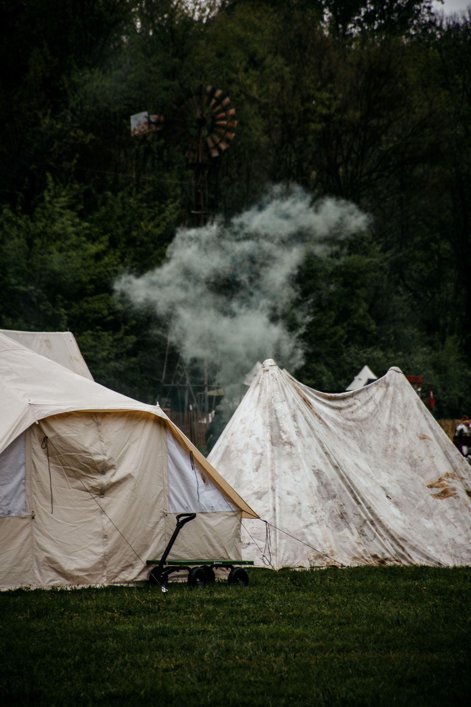White canvas tents set up on green grass at a campsite with smoke rising and a windmill visible among trees.