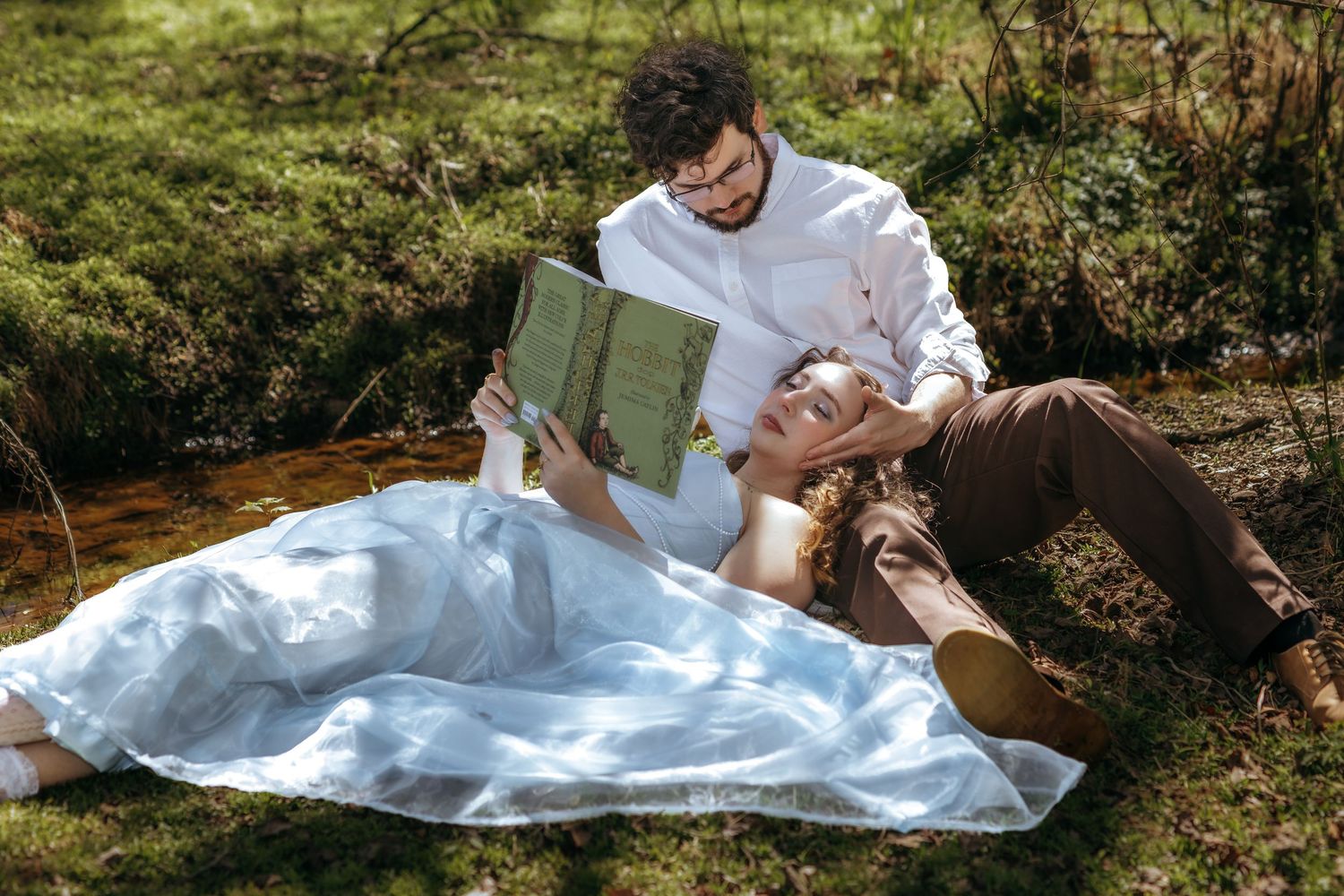 Couple relaxing outdoors on grass, woman lying down as man reads a book to her on a sunny day.