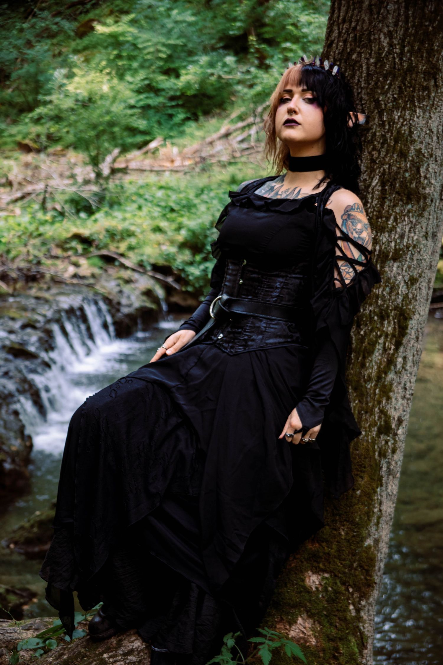 A figure in a flowing black dress reclines against a tree trunk beside a serene forest stream.