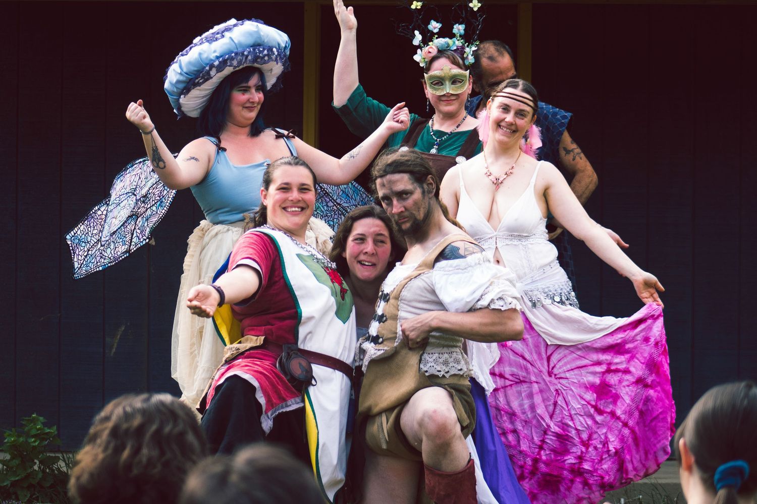 Performers in colorful costumes and period dress strike playful poses on stage during a theatrical production.