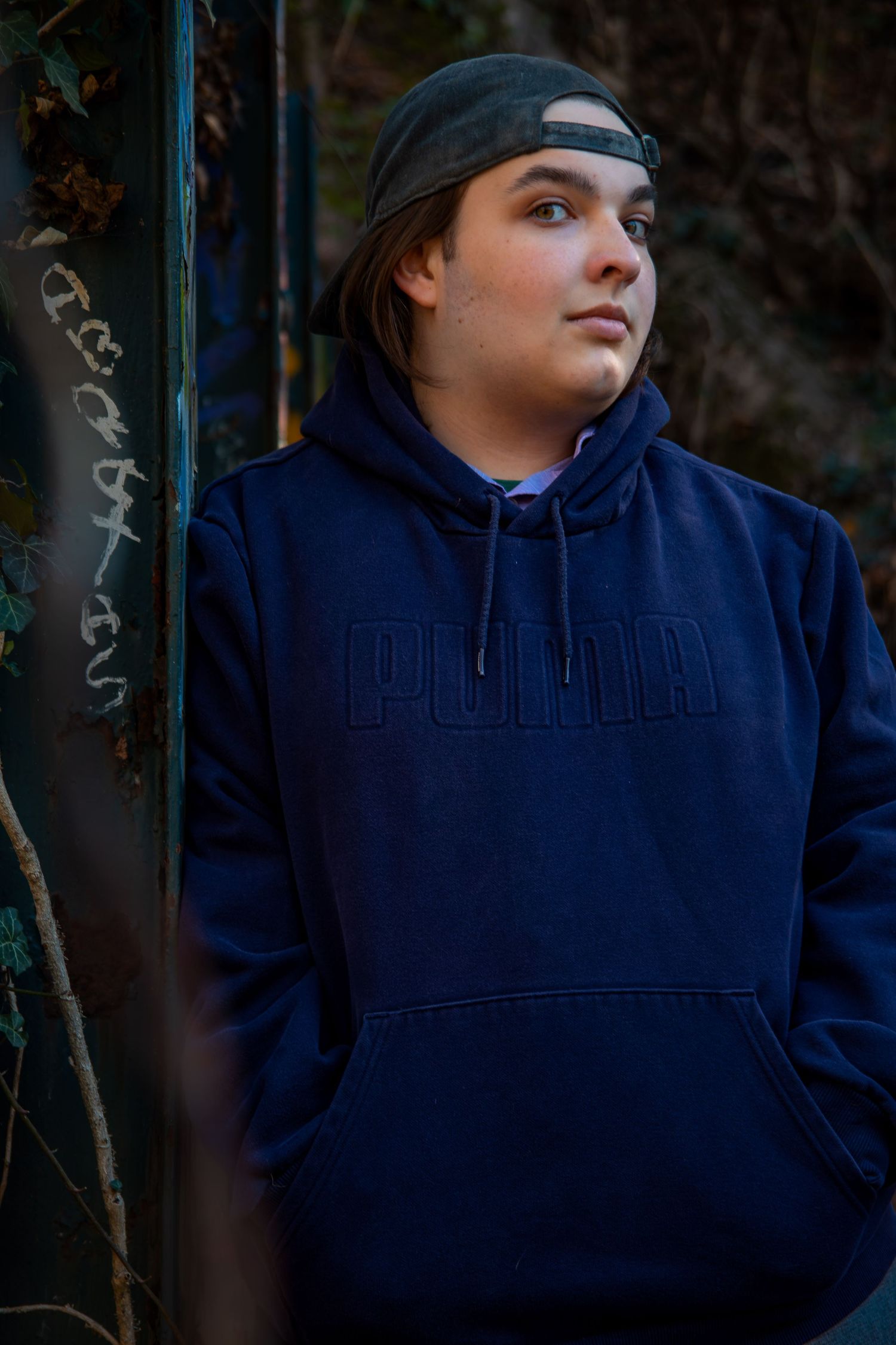Portrait of someone wearing a navy blue Puma hoodie and backwards baseball cap looking to the side against dark background.