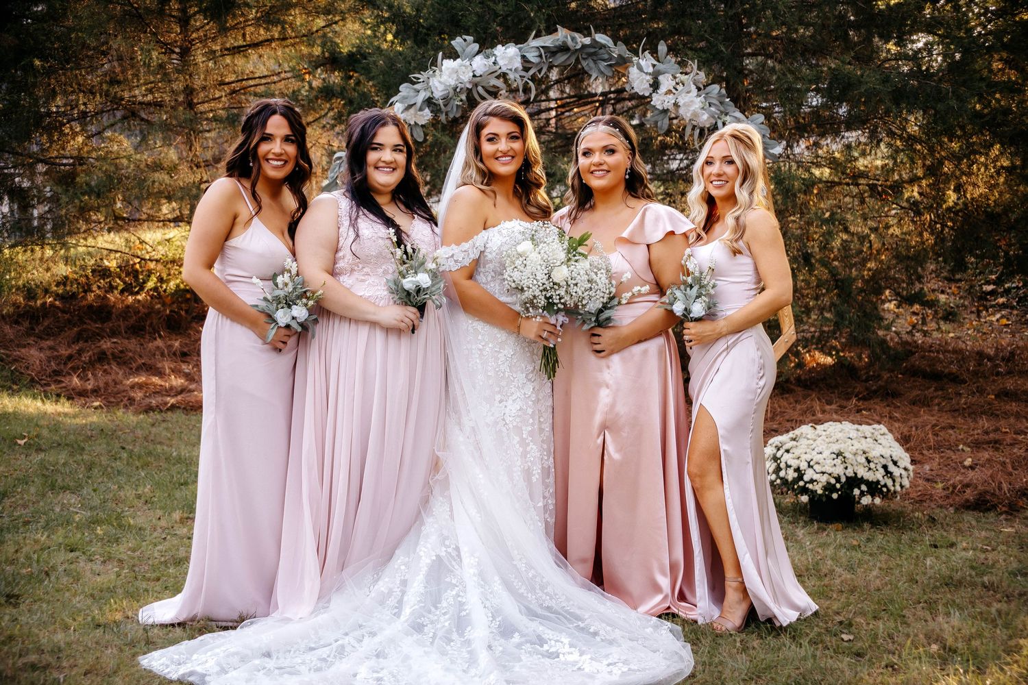 Wedding party in blush pink dresses poses together outdoors holding bouquets on a sunny autumn day.