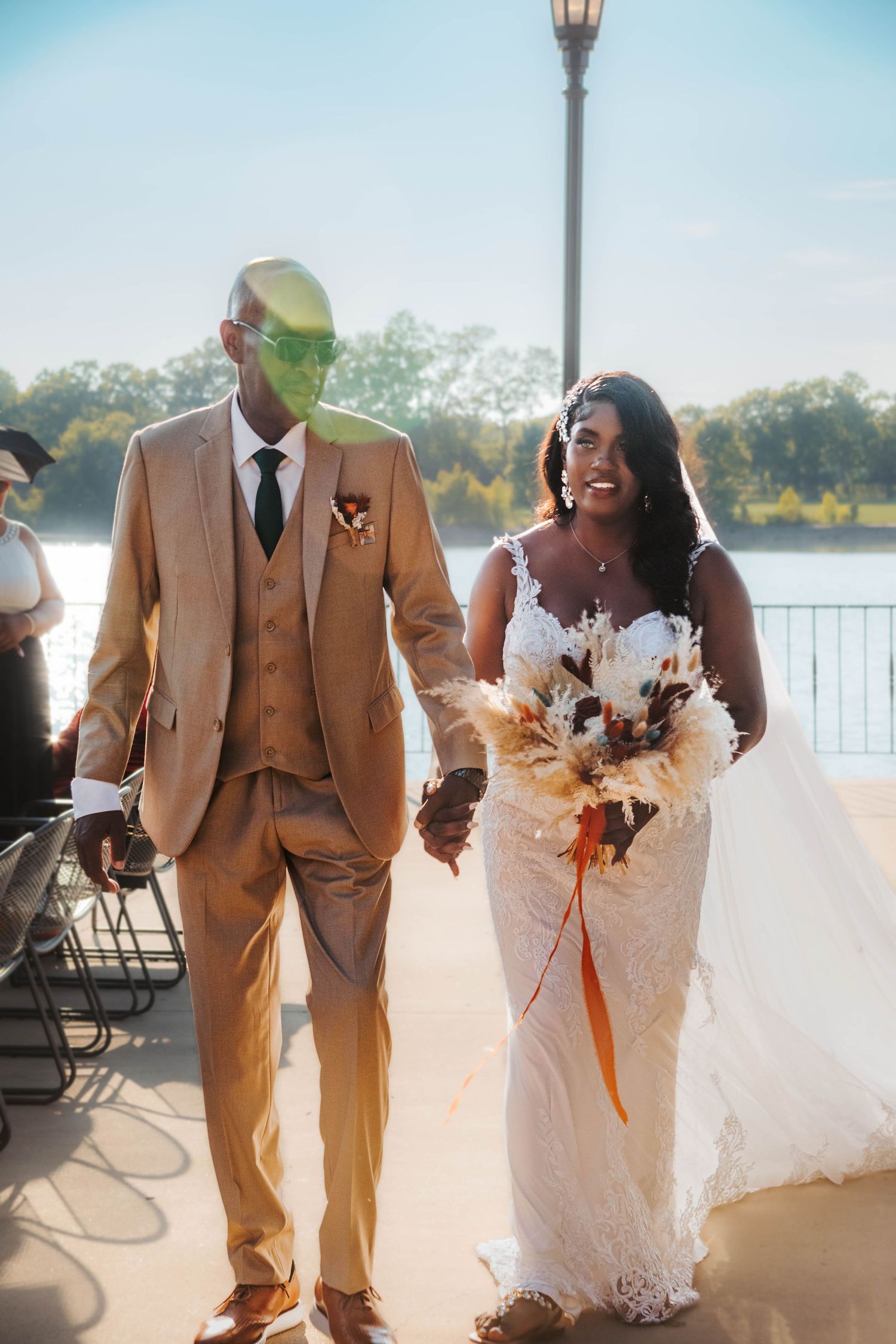 A wedding couple poses by a lakeside in matching tan and white formal attire with autumn-themed floral bouquets.