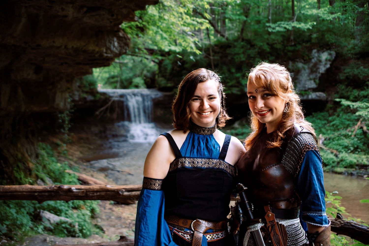 Two people in blue outfits explore a lush green forest trail with waterfalls and streams in the background.