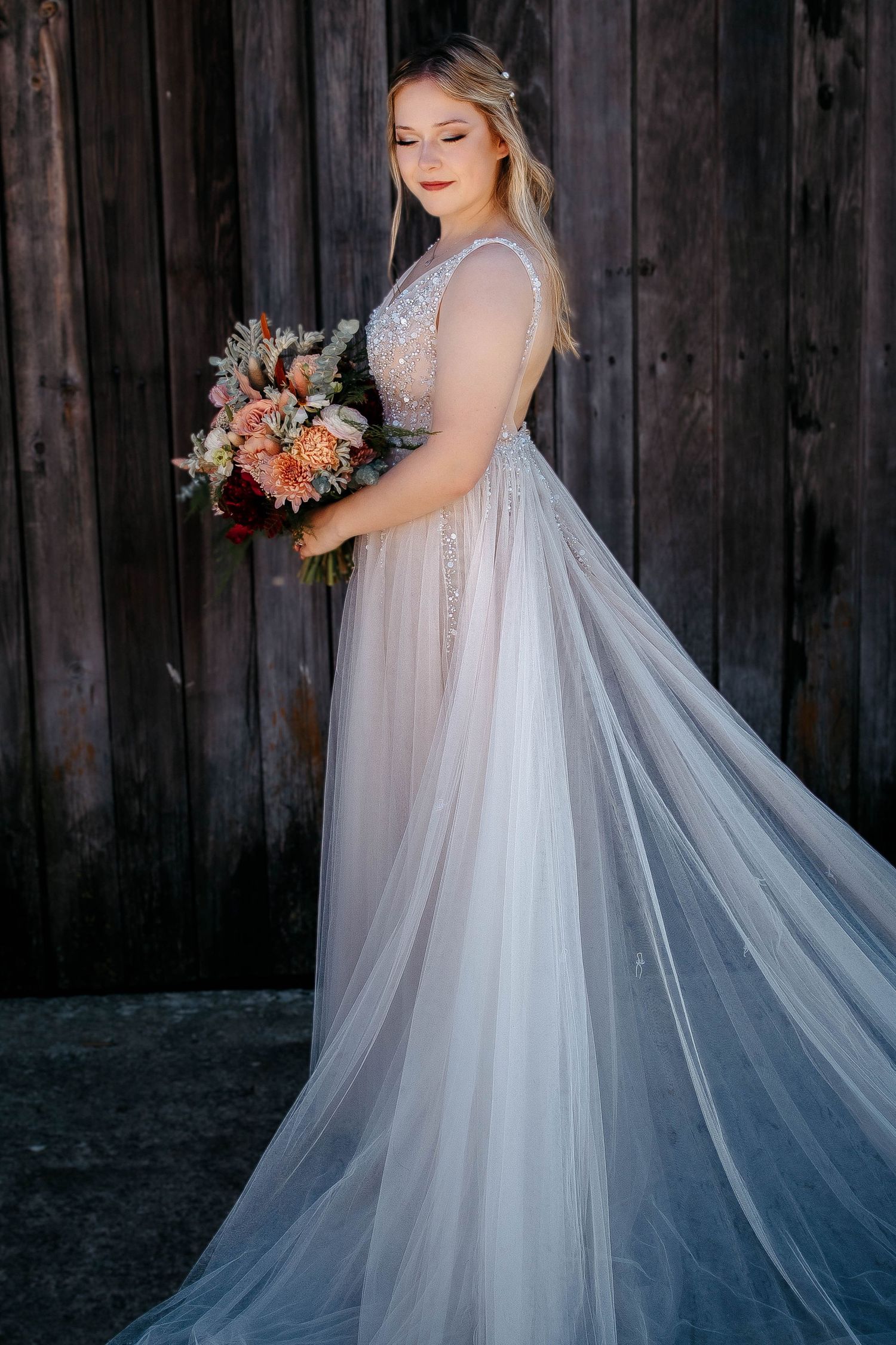 A bride in a flowing white lace gown holds an autumn-colored bouquet against a rustic wooden backdrop.