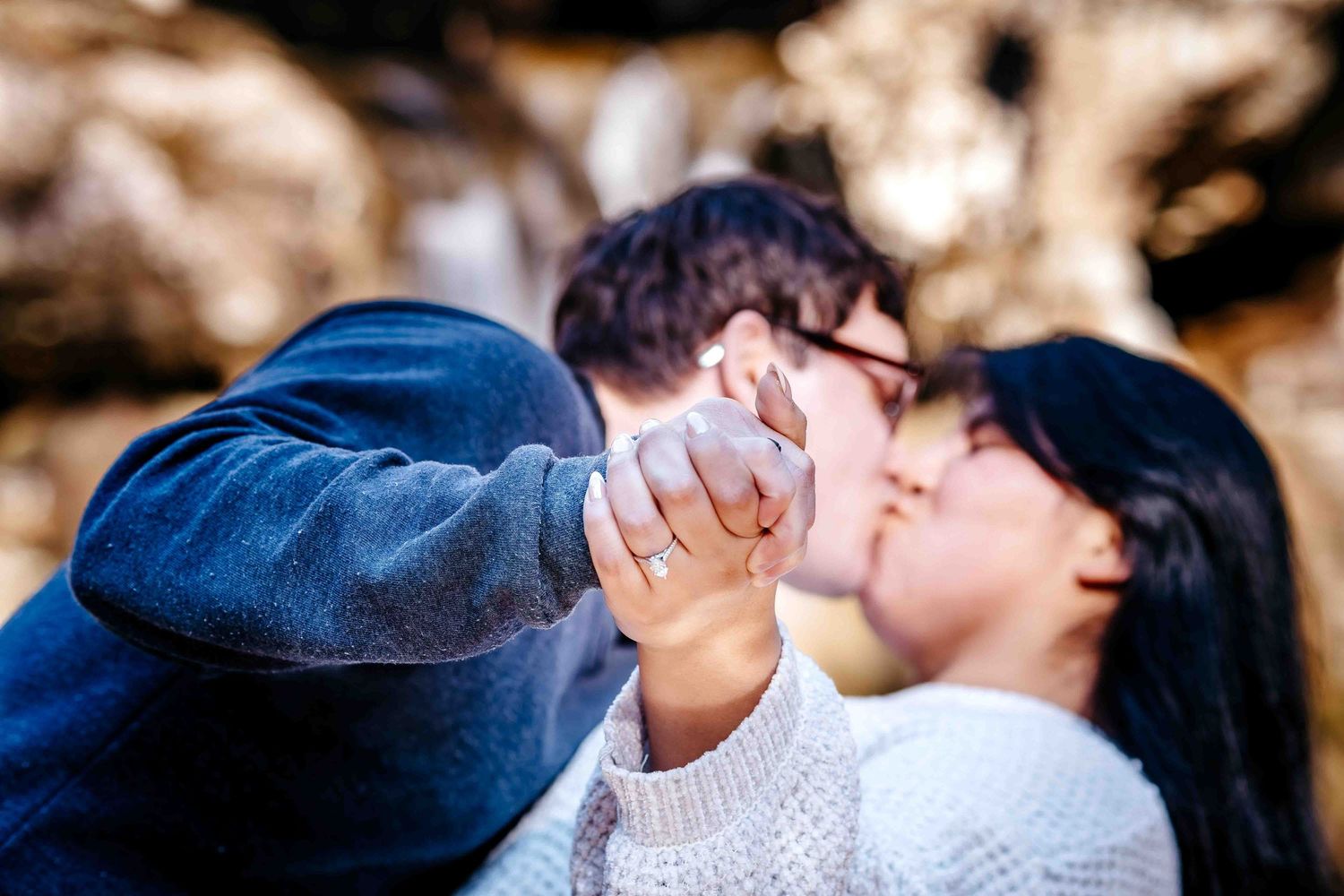 Romantic couple kissing while holding hands, engagement ring visible, soft bokeh background in warm tones.