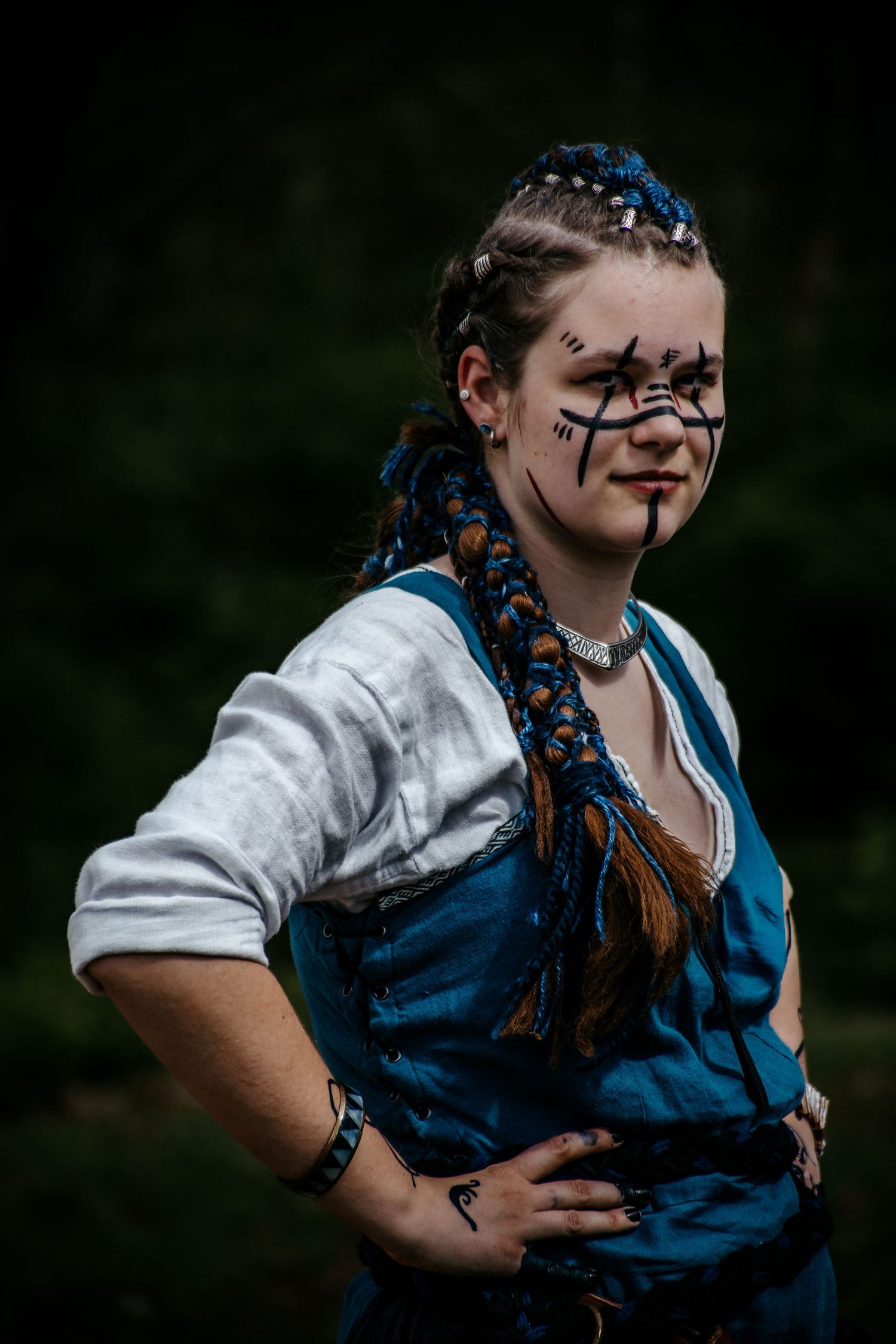 Woman in medieval costume with blue braids and face paint poses outdoors at a fantasy LARP event.