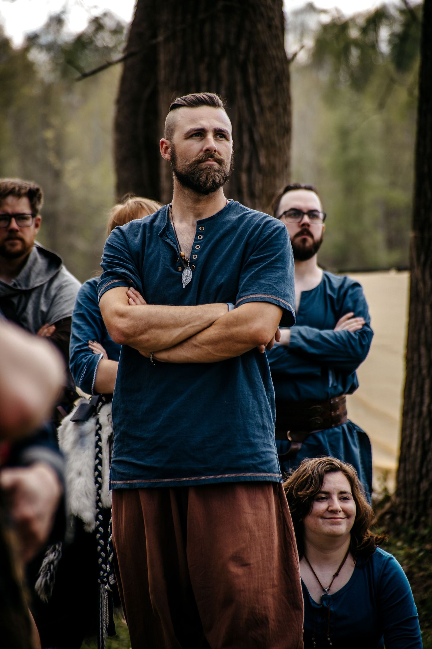 Bearded man in blue medieval tunic stands arms crossed outdoors at Renaissance faire event with group behind him.